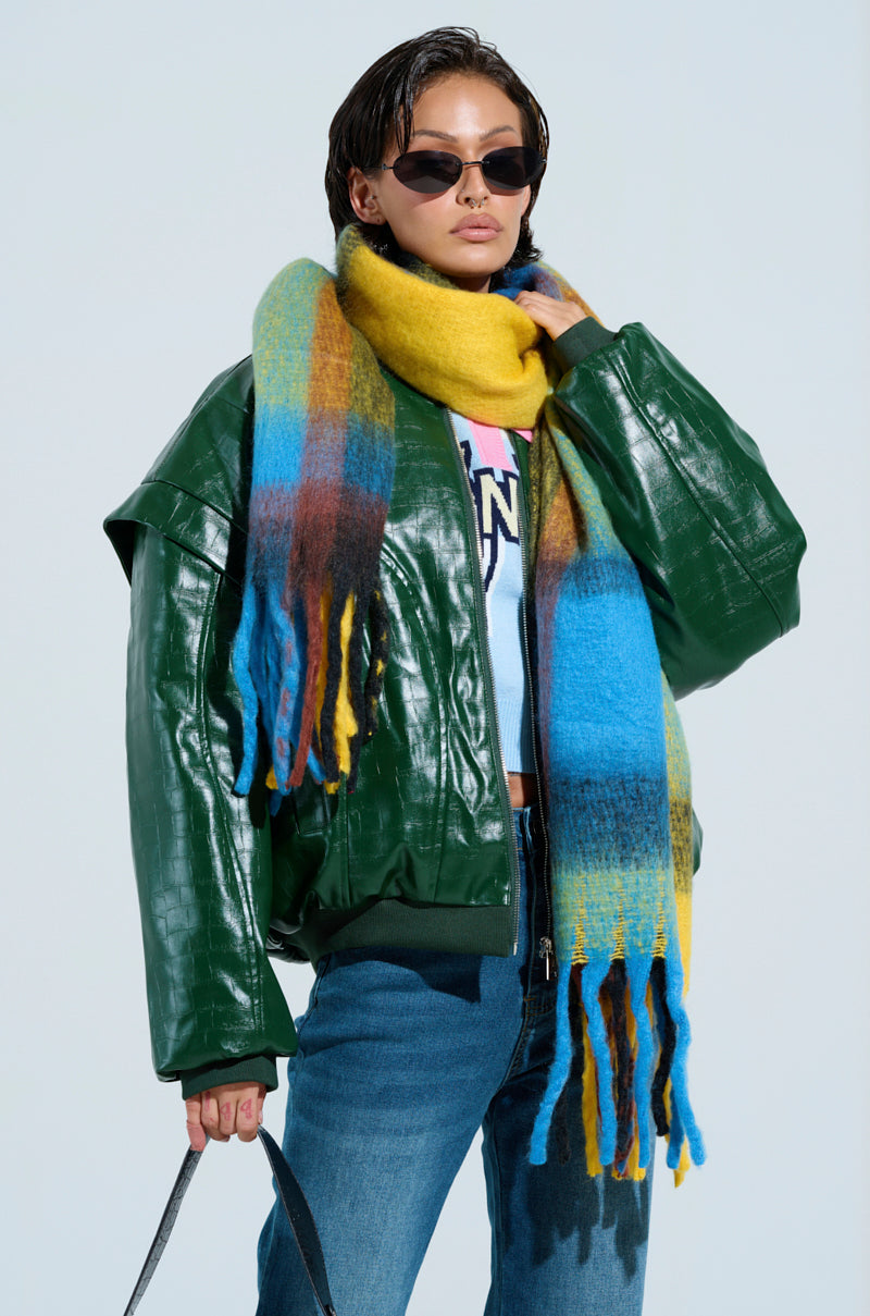 A person in a green leather jacket and blue jeans accessorizes with a cozy chic accessory, the oversized VARSITY PLAID SCARF. They're holding a small black bag, wearing black sunglasses against a light blue background.