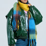 A person in a green leather jacket and blue jeans accessorizes with a cozy chic accessory, the oversized VARSITY PLAID SCARF. They're holding a small black bag, wearing black sunglasses against a light blue background.