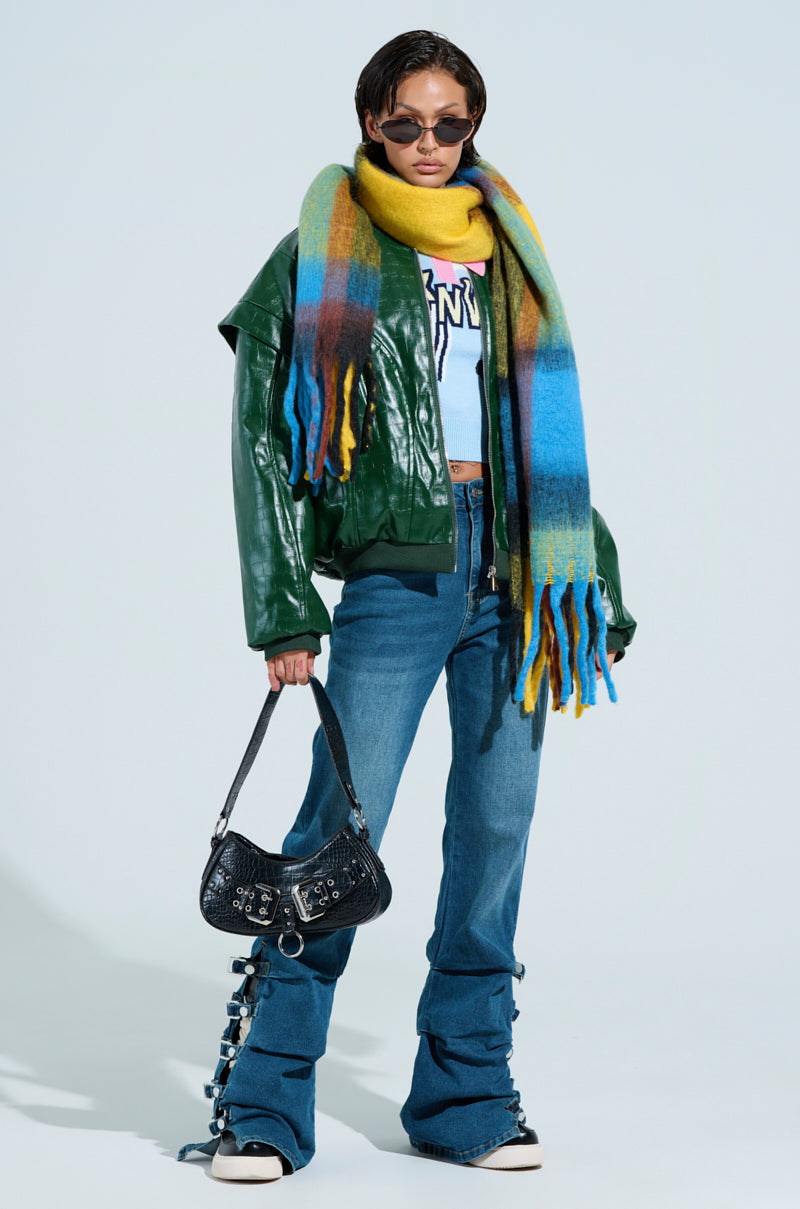 A person exudes cozy chic style with a large, VARSITY PLAID SCARF, green leather jacket, blue graphic sweater, wide-legged jeans, and black sneakers. They're holding a black handbag and wearing sunglasses against a light background.