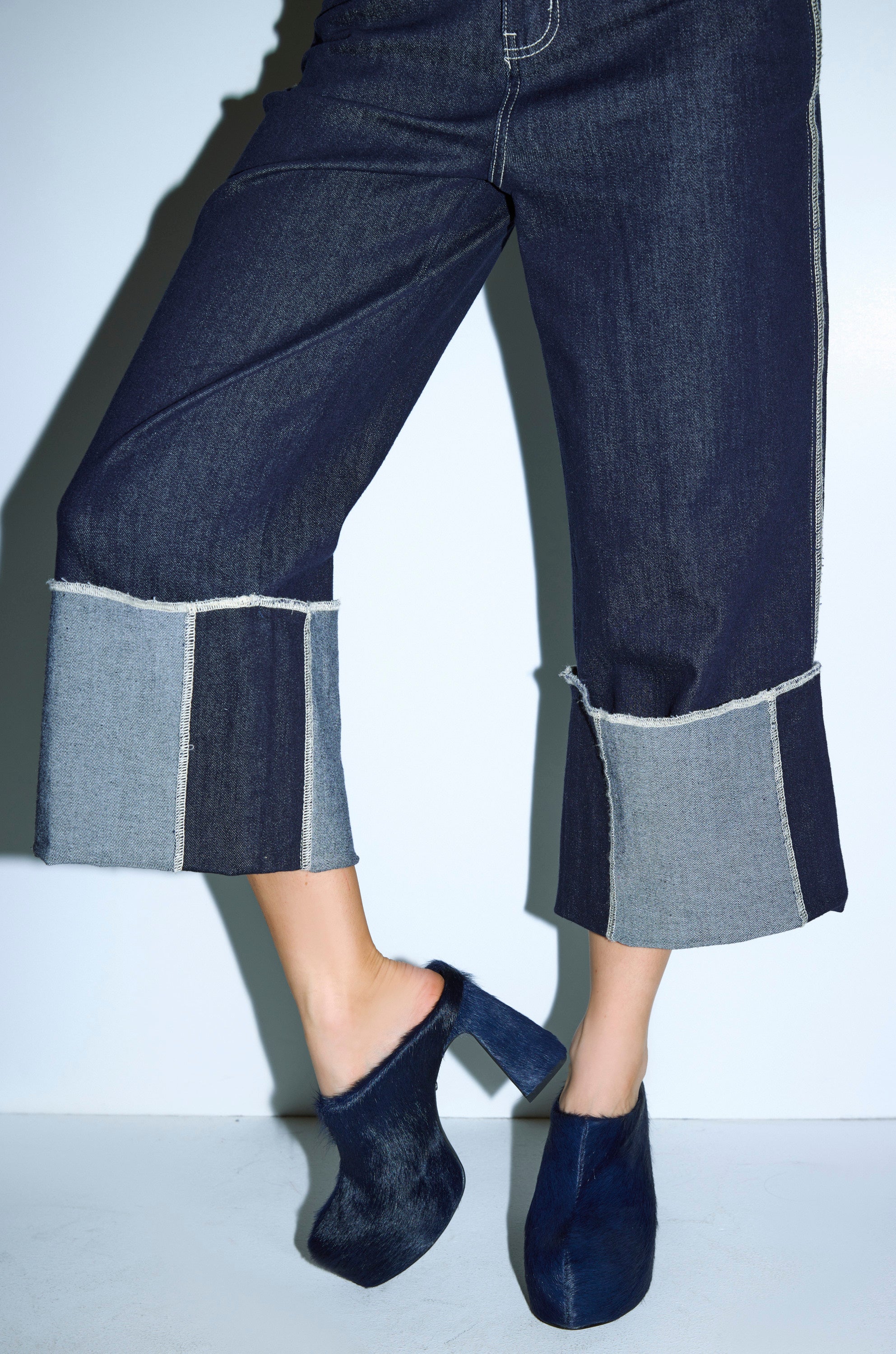 A person stands against a light background wearing wide-leg, dark denim jeans with large light cuffs and the AZALEA WANG GOLDING NAVY PONY HAIR CHUNKY PUMP. 0