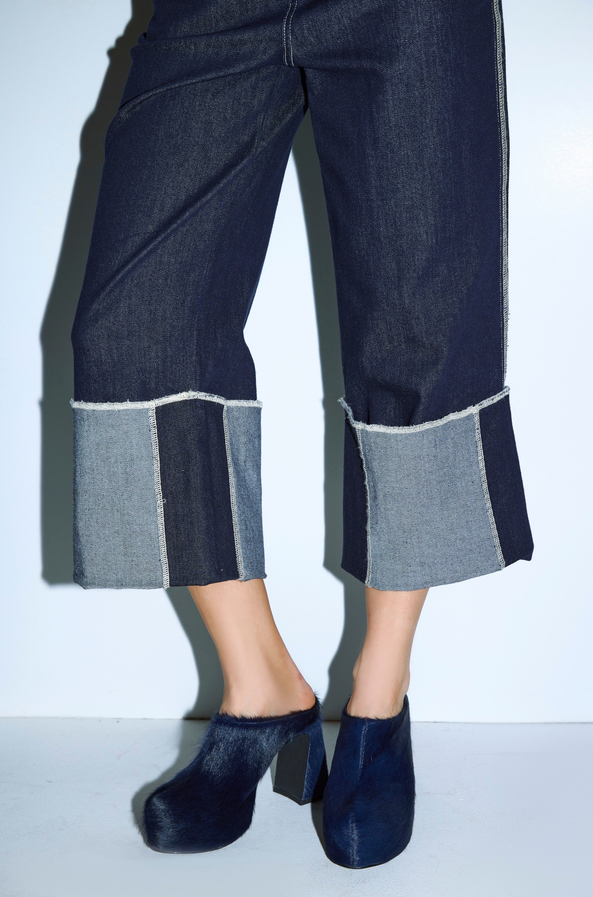 A person wears wide-leg cropped denim pants with contrasting hem panels, paired with AZALEA WANG GOLDING NAVY PONY HAIR CHUNKY PUMP heels. The background is a plain light color. 1