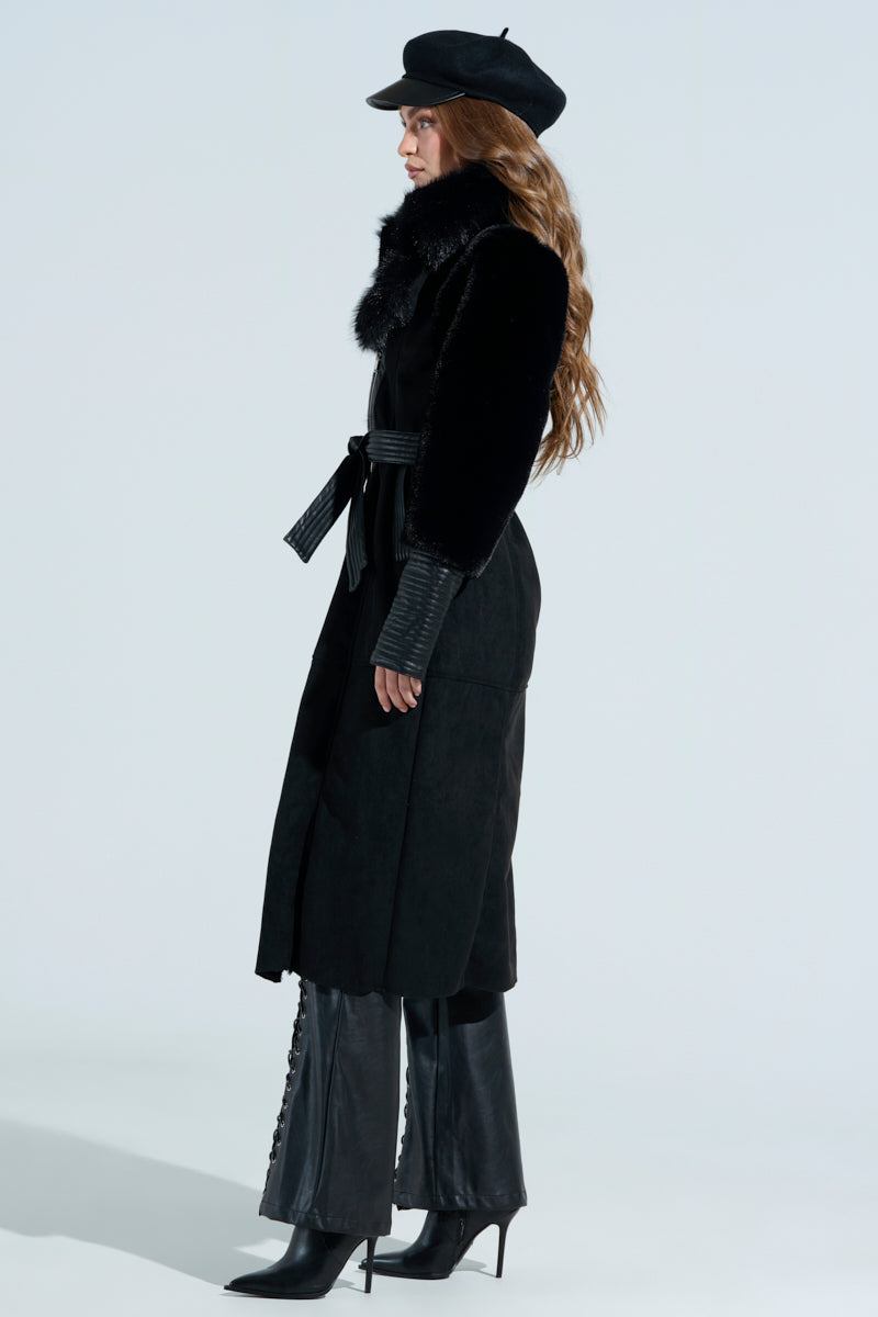 A person stands in profile against a plain background, wearing the GOODIE MIXED MEDIA COAT, a stylish matching hat, vegan leather pants, and high-heeled boots, with long hair cascading down their back.