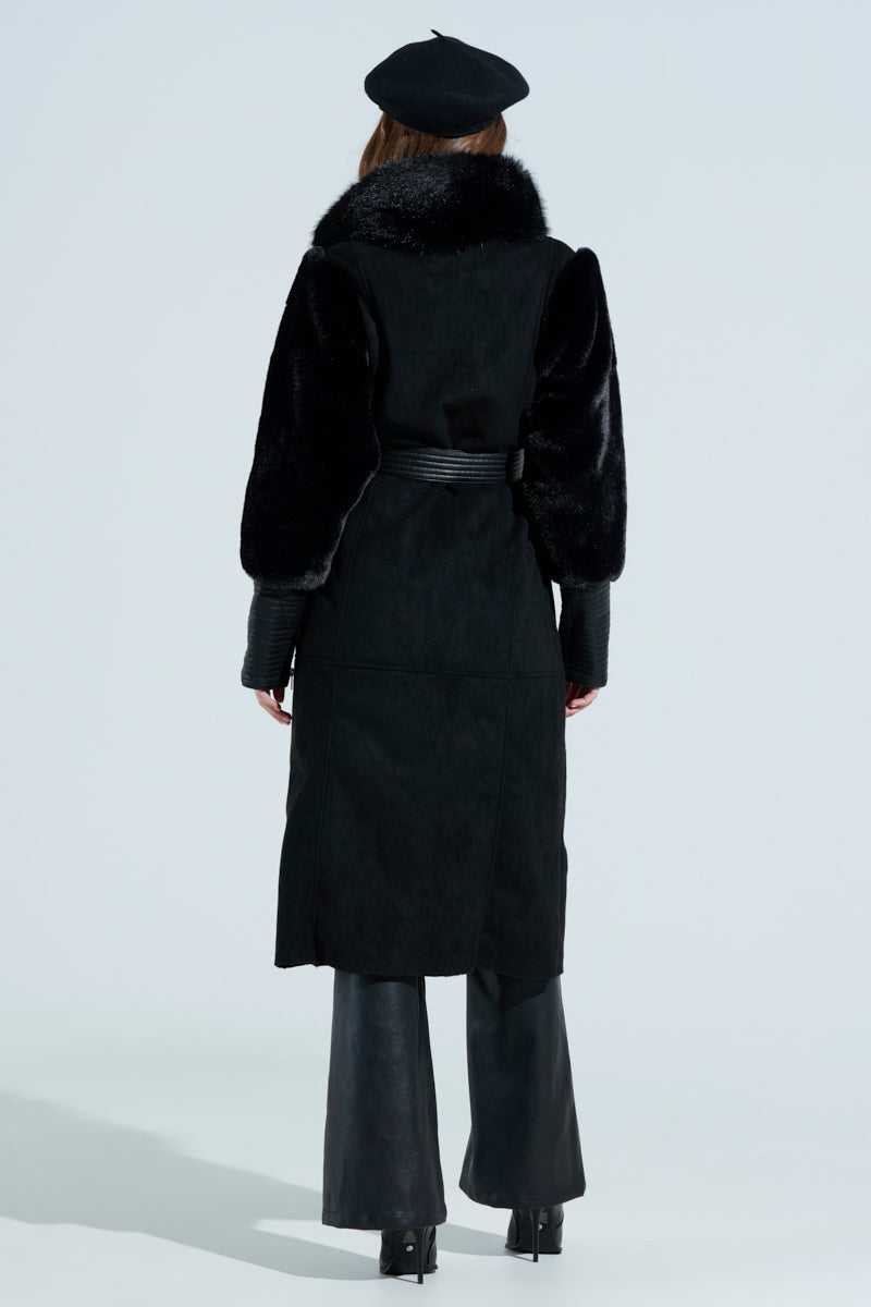 Identified from behind, the individual wears a GOODIE MIXED MEDIA COAT in black faux fur with textured sleeves and a cinched waist, complemented by a beret, wide-leg black pants, and high-heeled boots. The plain background enhances their stylish appearance.