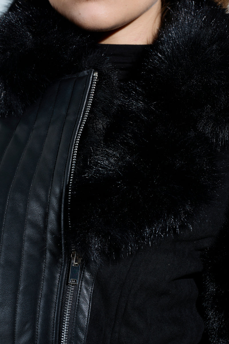 Close-up of a person in the GOODIE MIXED MEDIA COAT, featuring black vegan leather and a shiny, textured faux fur collar. The visible zipper and quilted details enhance its appeal, while lighting highlights the soft texture of the fur.