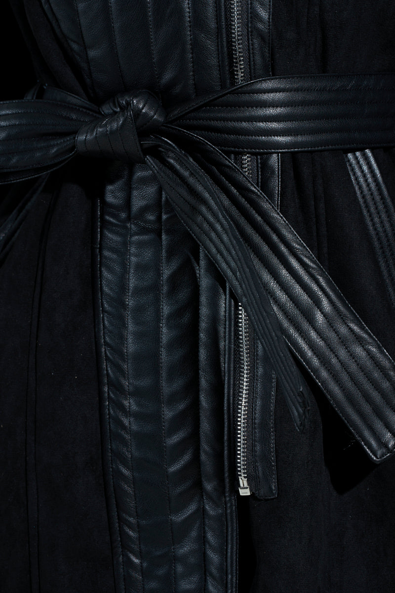 Close-up of the GOODIE MIXED MEDIA COAT in black vegan leather, featuring a quilted texture. A leather belt loosely knots at the front, revealing a metal zipper below. The material is smooth and shiny.