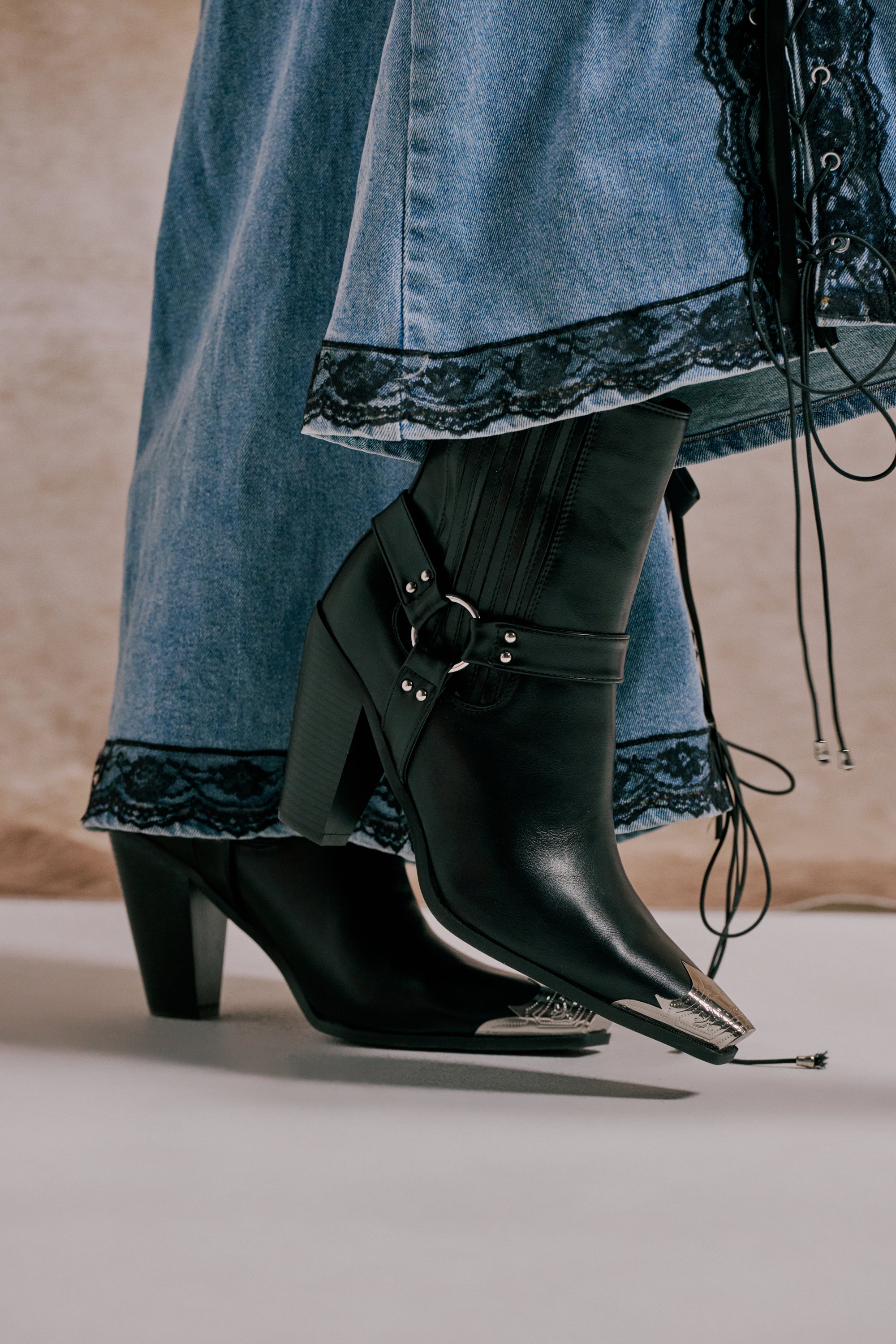 Close-up of a person wearing the AZALEA WANG GOODMAN BLACK CHUNKY WESTERN HARDWARE PU BOOTIE with a long blue denim skirt featuring black lace trim and corset-style laces on the side. 0