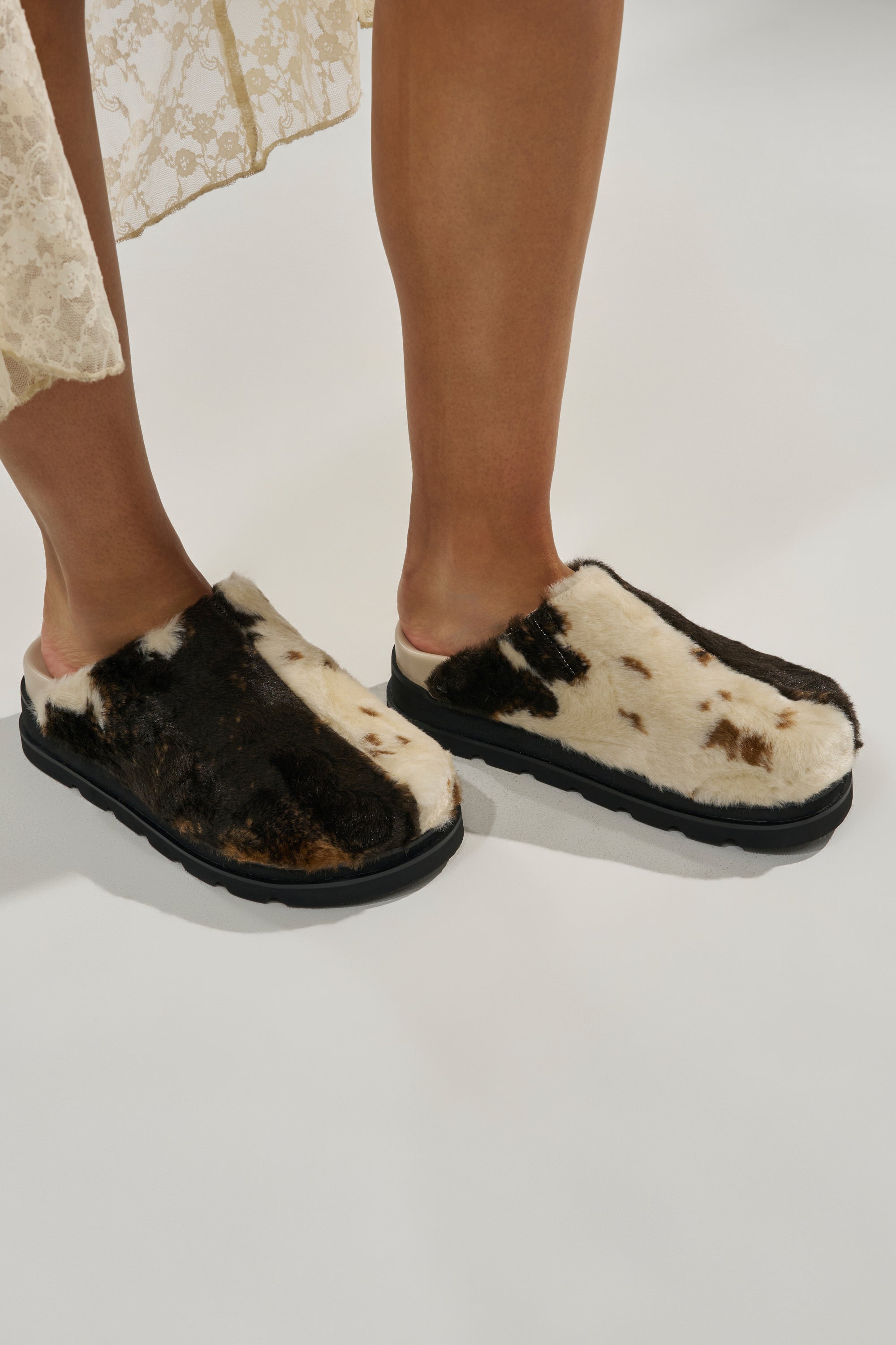 A person wearing the AZALEA WANG GOVINDA MULTI SLIP ON COW PRINT CLOG and a cream lace skirt stands on a white surface.
