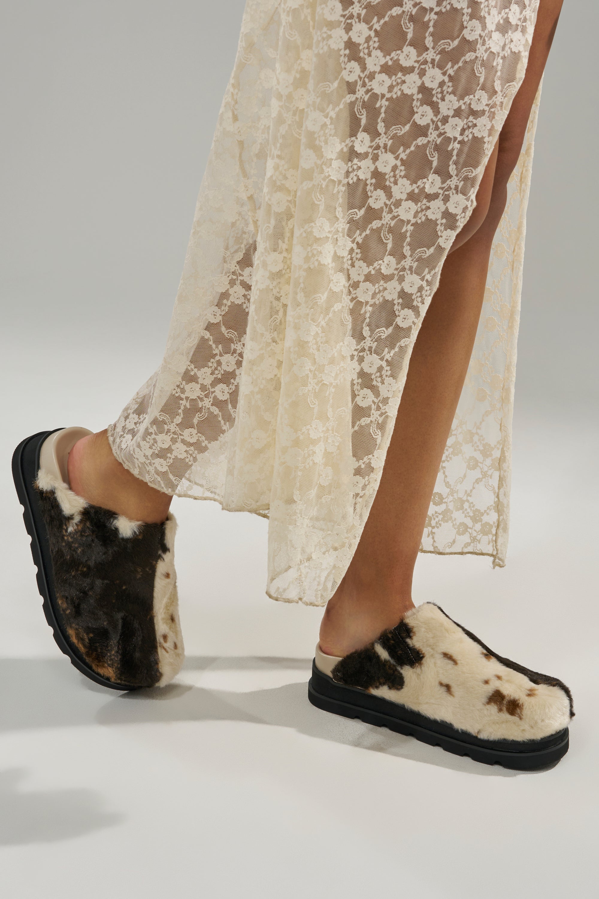 A person in a sheer cream lace dress wears AZALEA WANG GOVINDA MULTI SLIP ON COW PRINT CLOGS, standing on a light gray background. Only their legs and feet are shown.