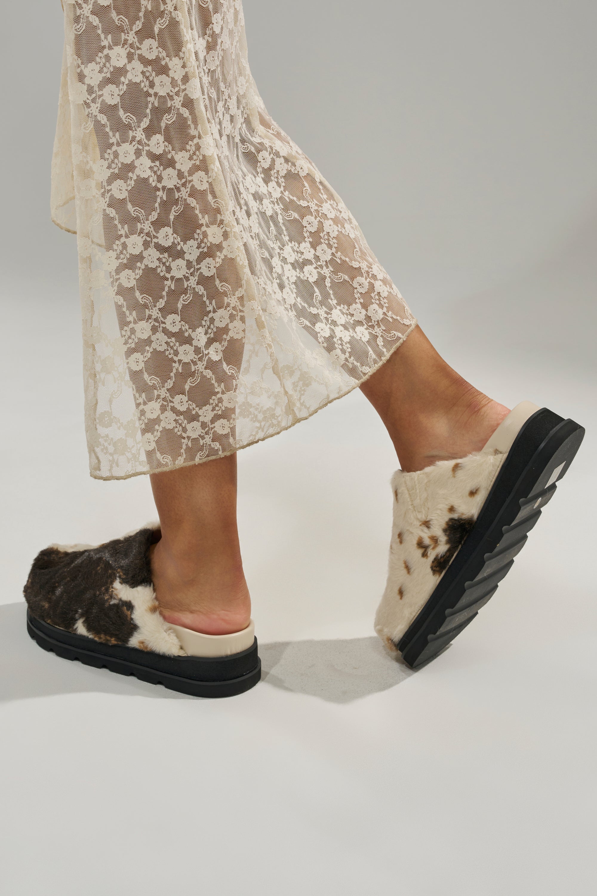 A person wearing a long sheer lace skirt and the AZALEA WANG GOVINDA MULTI SLIP ON COW PRINT CLOG stands on a light gray background.