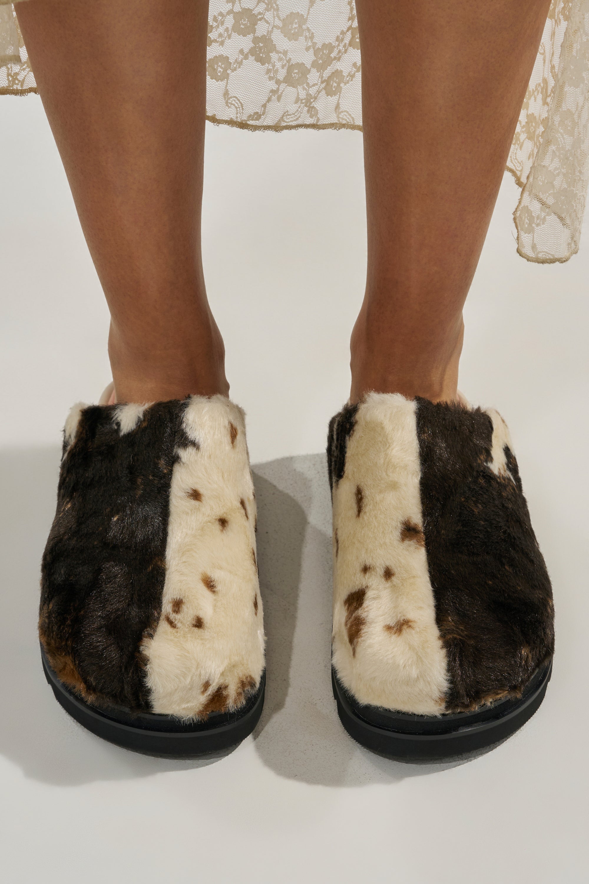 A person wearing the AZALEA WANG GOVINDA MULTI SLIP ON COW PRINT CLOG stands on a white surface. The cozy clogs feature a fuzzy cow print with a black sole, paired with a beige lace skirt.