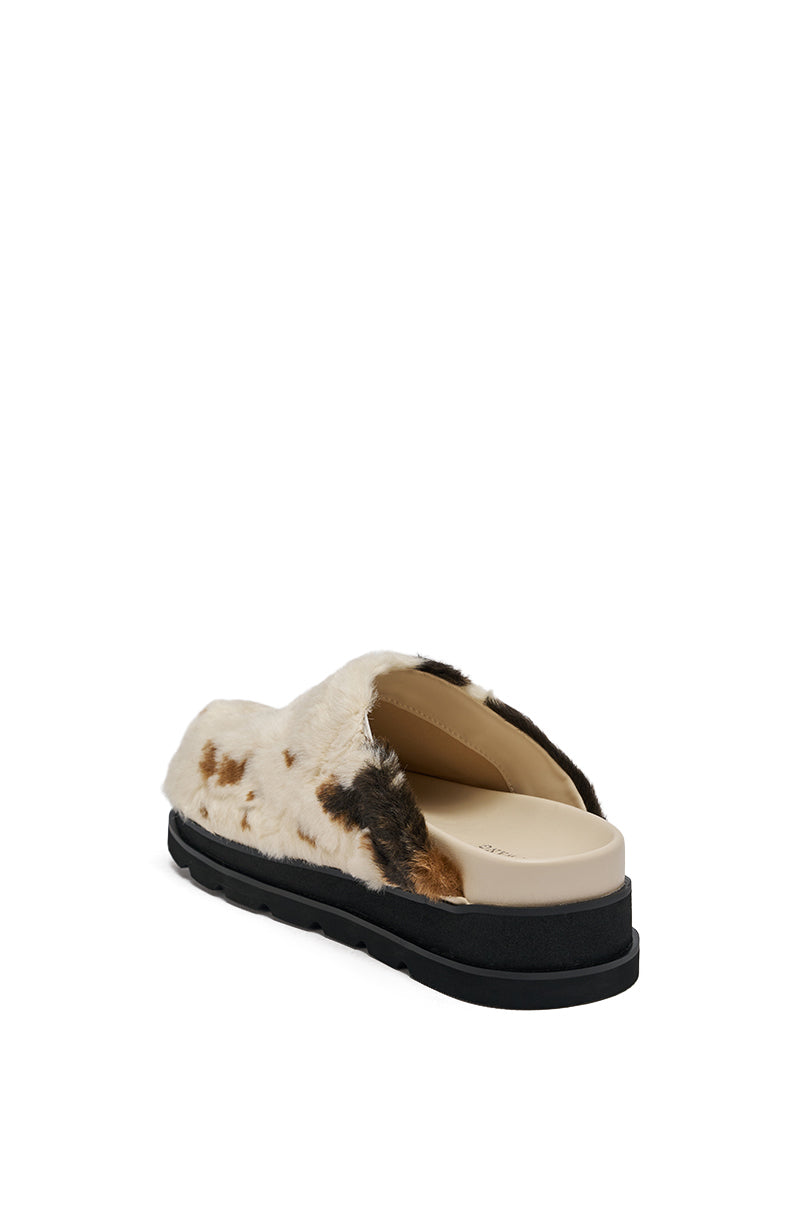 The AZALEA WANG GOVINDA MULTI SLIP ON COW PRINT CLOG features a fuzzy cow-print design, faux fur, open heel, tan insole, and thick black sole, shown angled from the back on a white background.