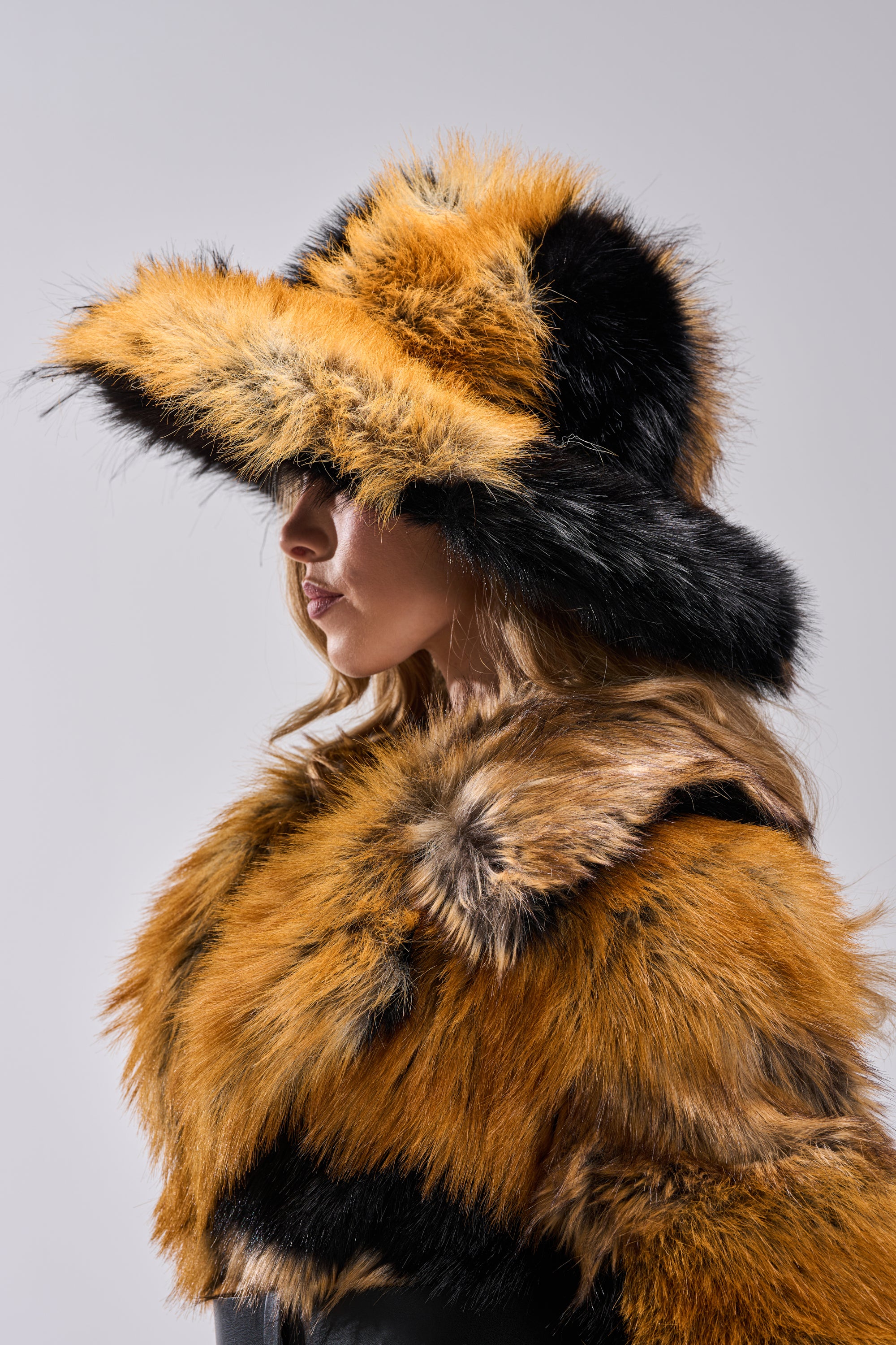 A woman in profile wears the COSMO FAUX FUR OVERSIZED BUCKET HAT and a multicolored faux fur coat against a plain gray background. The brim shadows her face, drawing attention to this bold winter accessory.