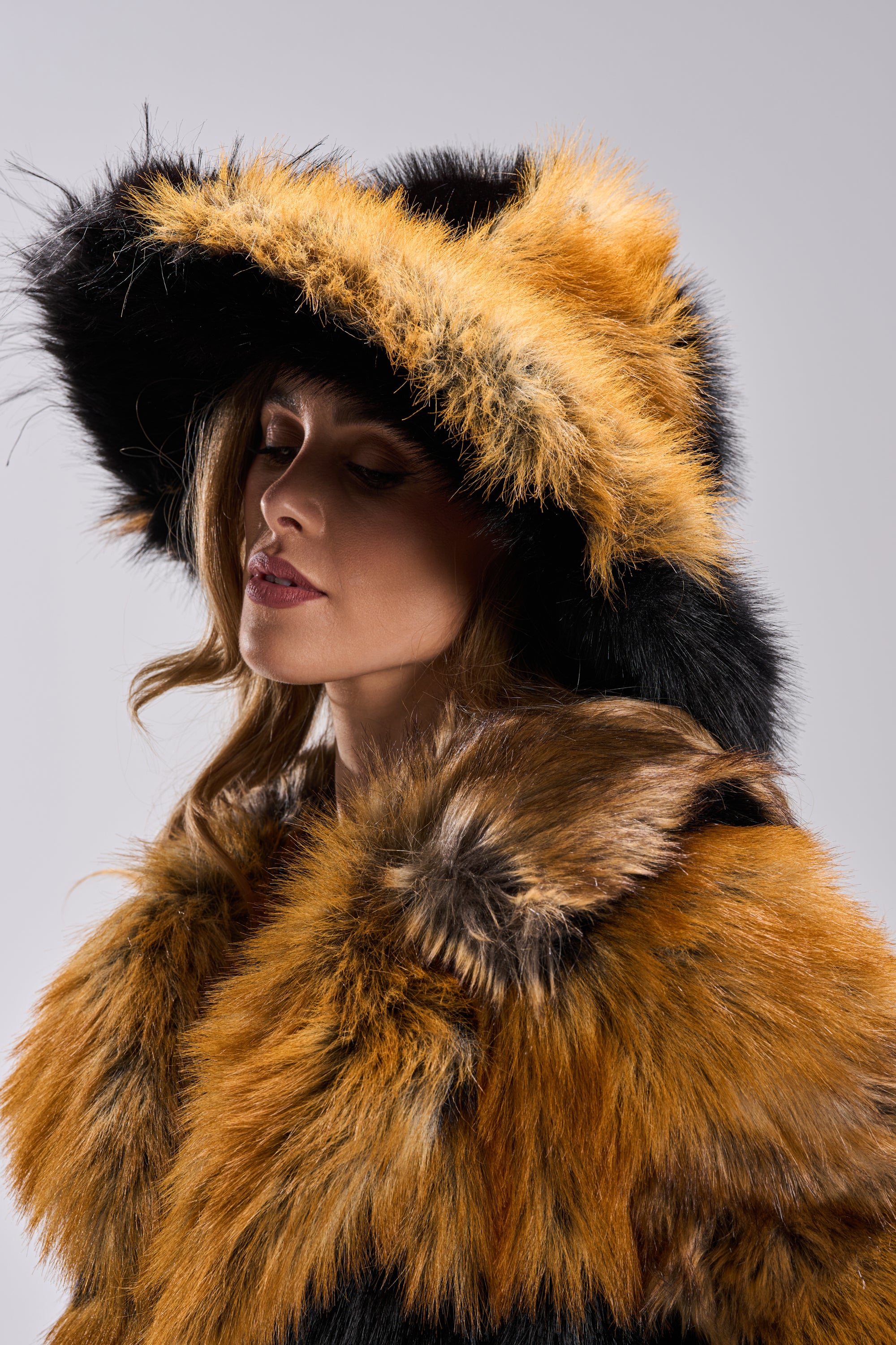 A woman with light wavy hair stands against a light gray background wearing the COSMO FAUX FUR OVERSIZED BUCKET HAT and a matching coat in black, orange, and brown—a bold winter accessory statement with her eyes closed.