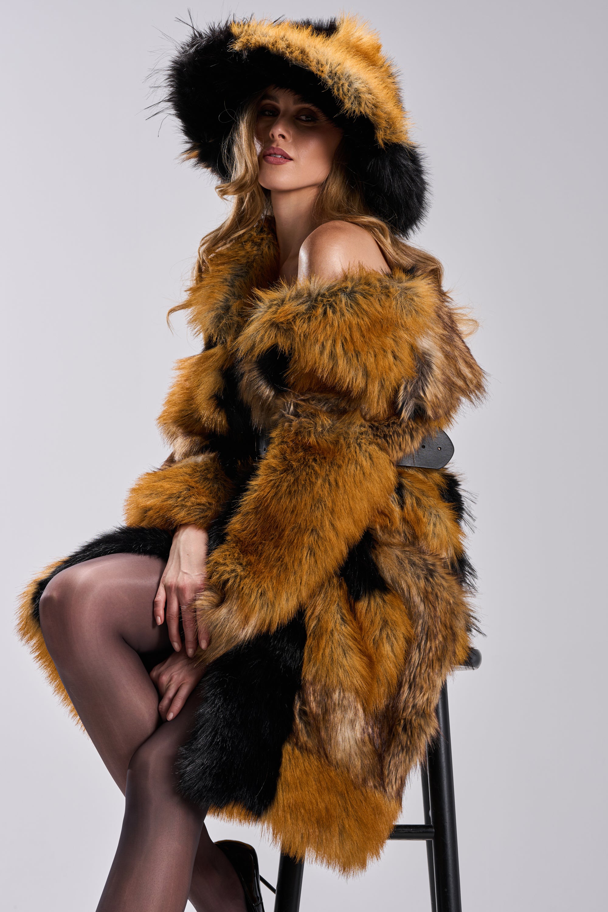 A woman with wavy hair confidently showcases the COSMO FAUX FUR OVERSIZED BUCKET HAT, pairing it with a bold orange and black fur coat as she sits on a stool and gazes to the side.