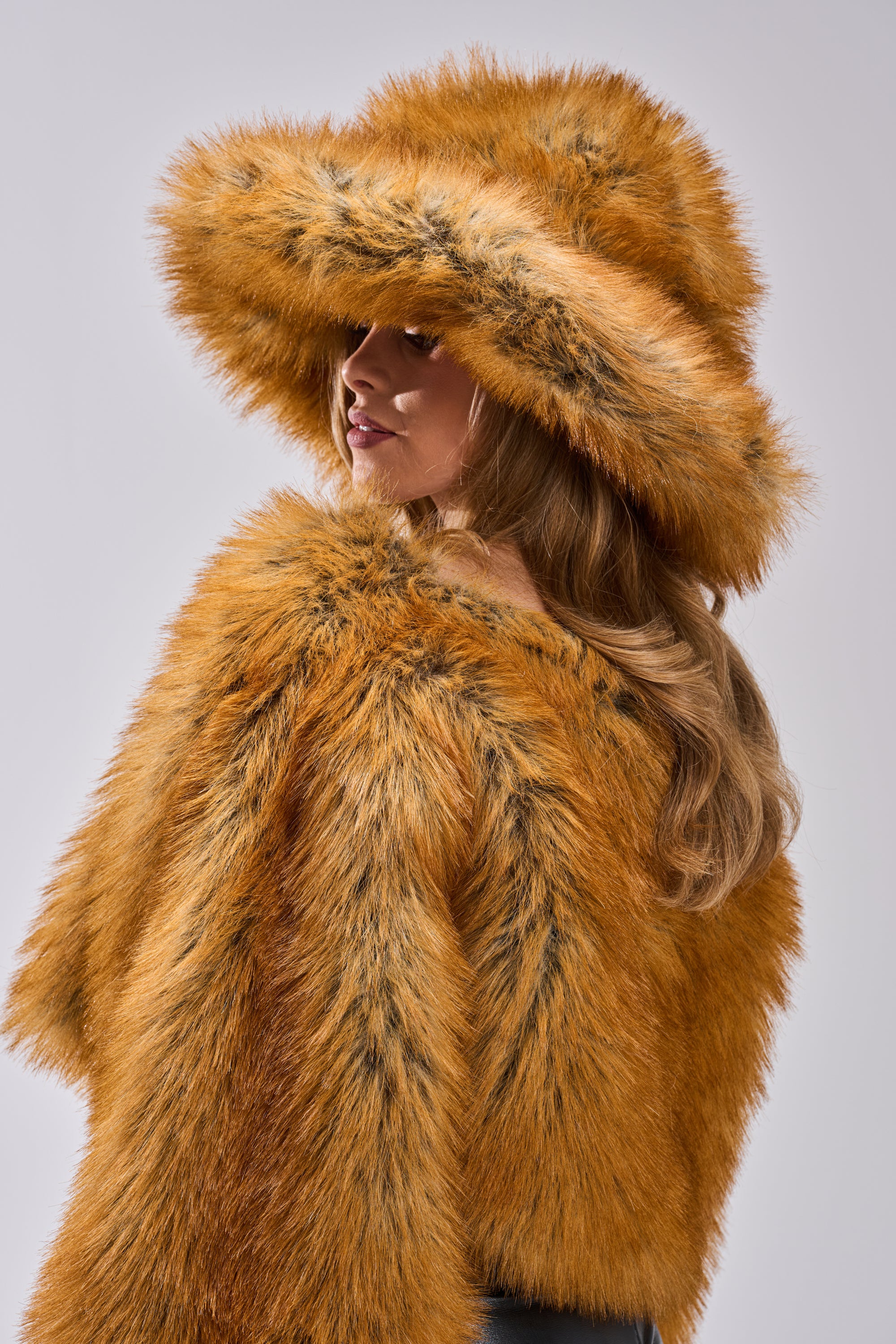 A woman with long wavy hair wears the HOT GIRL FAUX FUR OVERSIZED BUCKET HAT in bold orange, paired with a matching faux fur coat, as she stands against a neutral grey background.