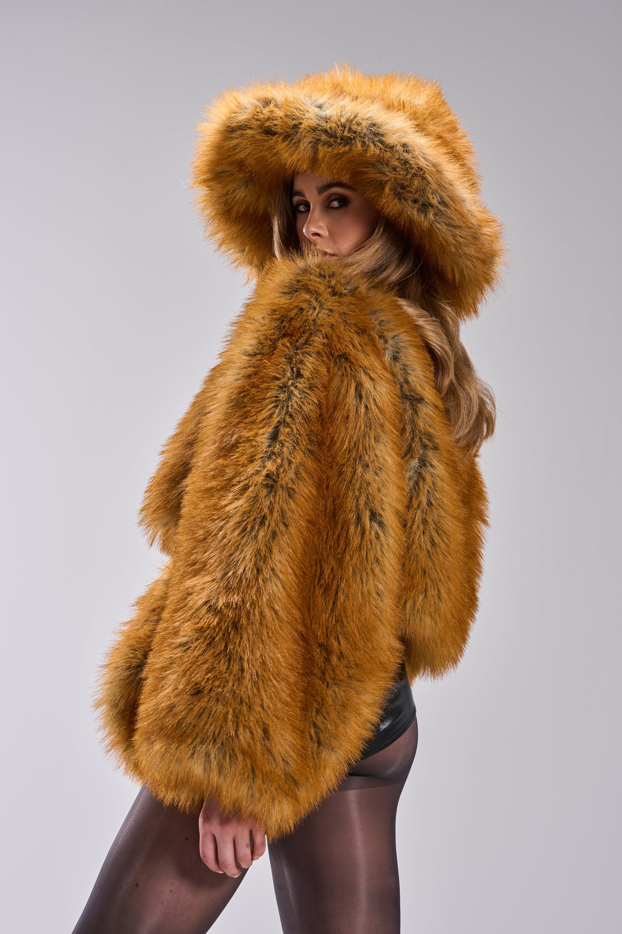 A woman in a large orange faux fur coat and long hair glances back, styling black shorts and sheer tights with the HOT GIRL FAUX FUR OVERSIZED BUCKET HAT, set against a plain light background.