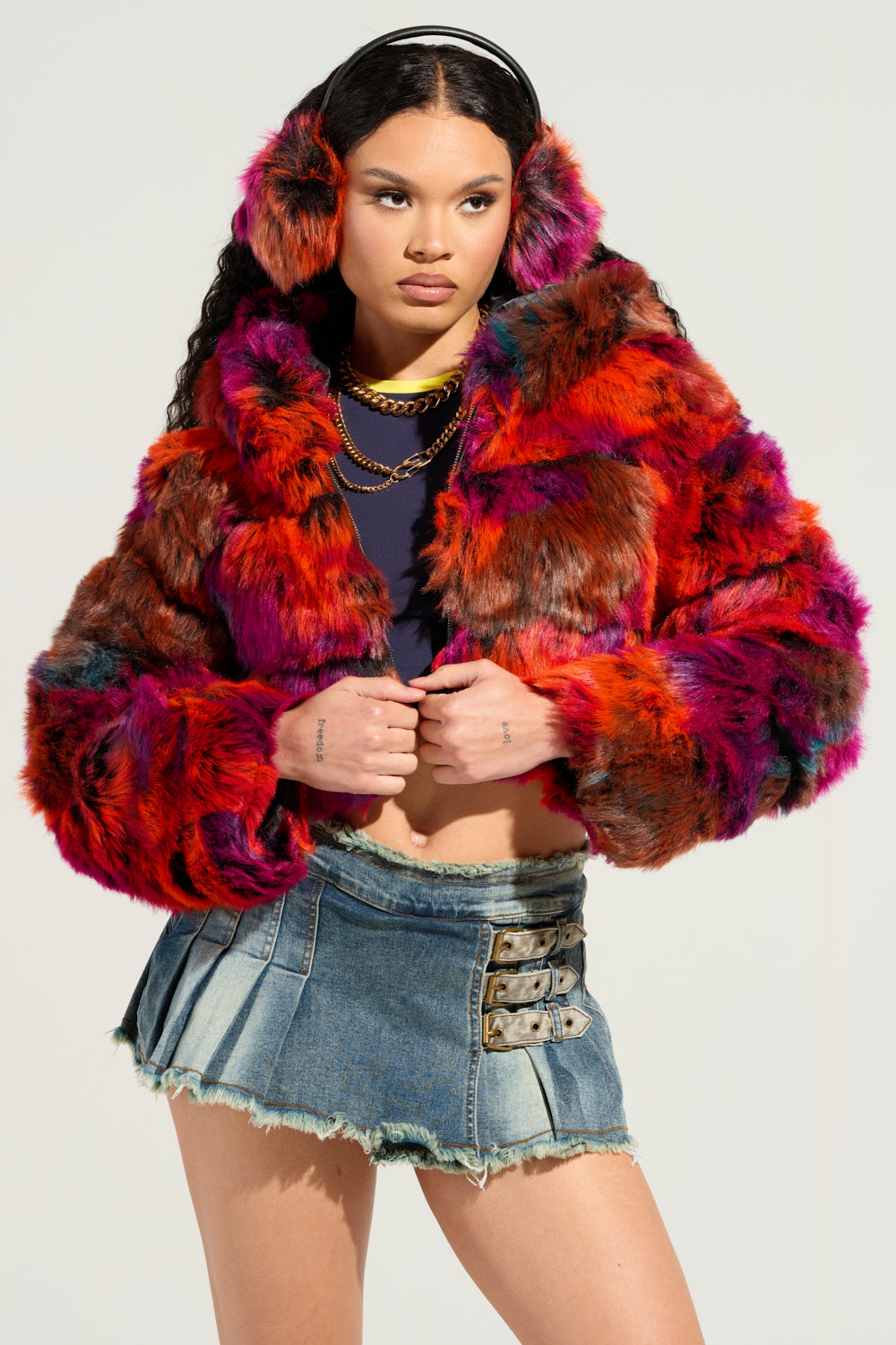 A person flaunts the GRACELLE MULTI FAUX FUR CROPPED JACKET, accompanied by matching earmuffs and a chain necklace. They boast long curly hair and rock a denim mini skirt with buckle details. The plain background beautifully highlights their bold fashion statement. 2
