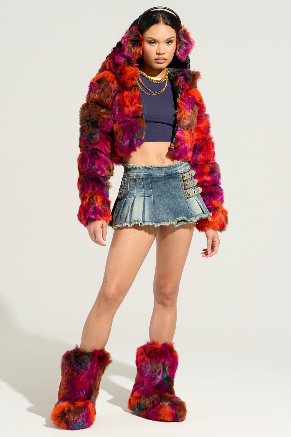 A person confidently stands out in the GRACELLE MULTI FAUX FUR CROPPED JACKET, paired with matching earmuffs and boots. They complement the look with a dark crop top and a denim mini skirt featuring buckle details, while the plain background accentuates their striking outfit. 10