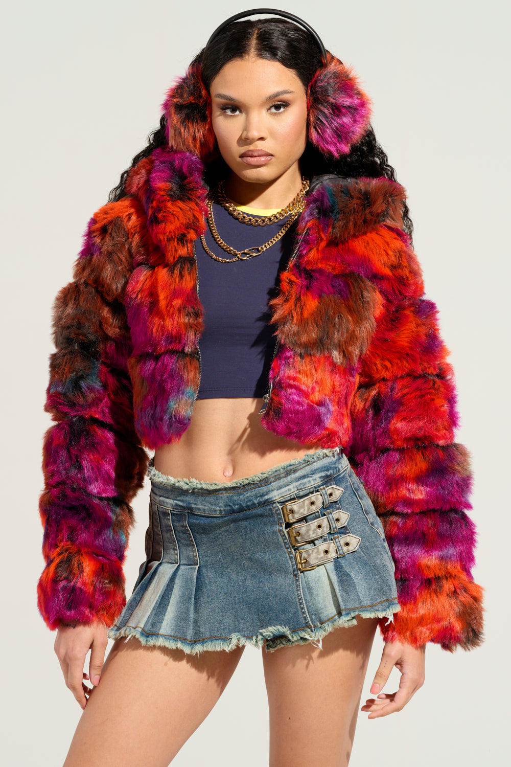 A person styled in the GRACELLE MULTI FAUX FUR CROPPED JACKET, paired with matching earmuffs, a dark crop top, and a denim mini skirt with buckle details. This ensemble effortlessly combines bold textures and colors. 3