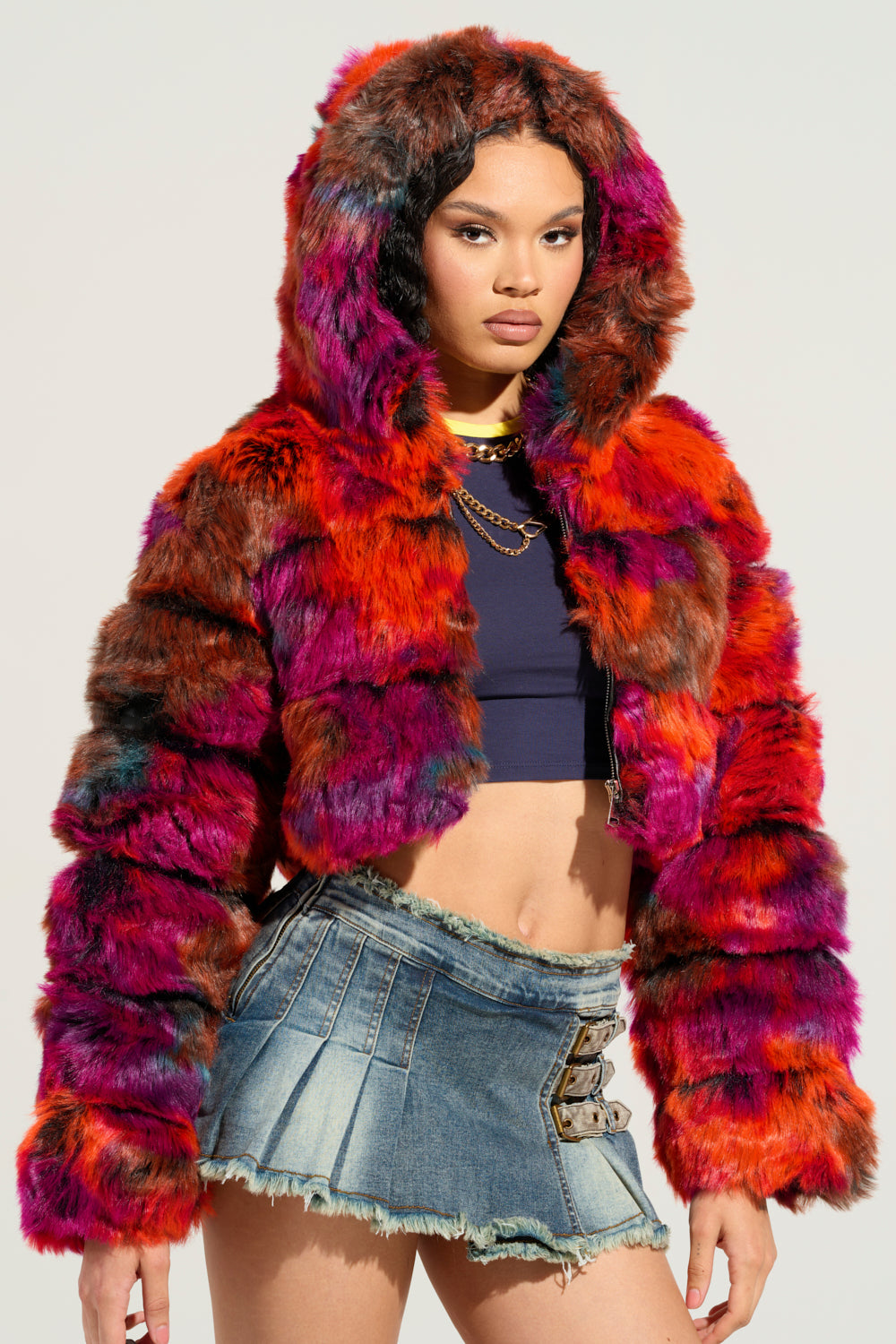 A person pairs the GRACELLE MULTI FAUX FUR CROPPED JACKET, known for its vibrant, multicolored design and hood, with a black crop top and a denim mini skirt that features buckle details. The plain light-colored backdrop accentuates the standout multi-color jacket. 0
