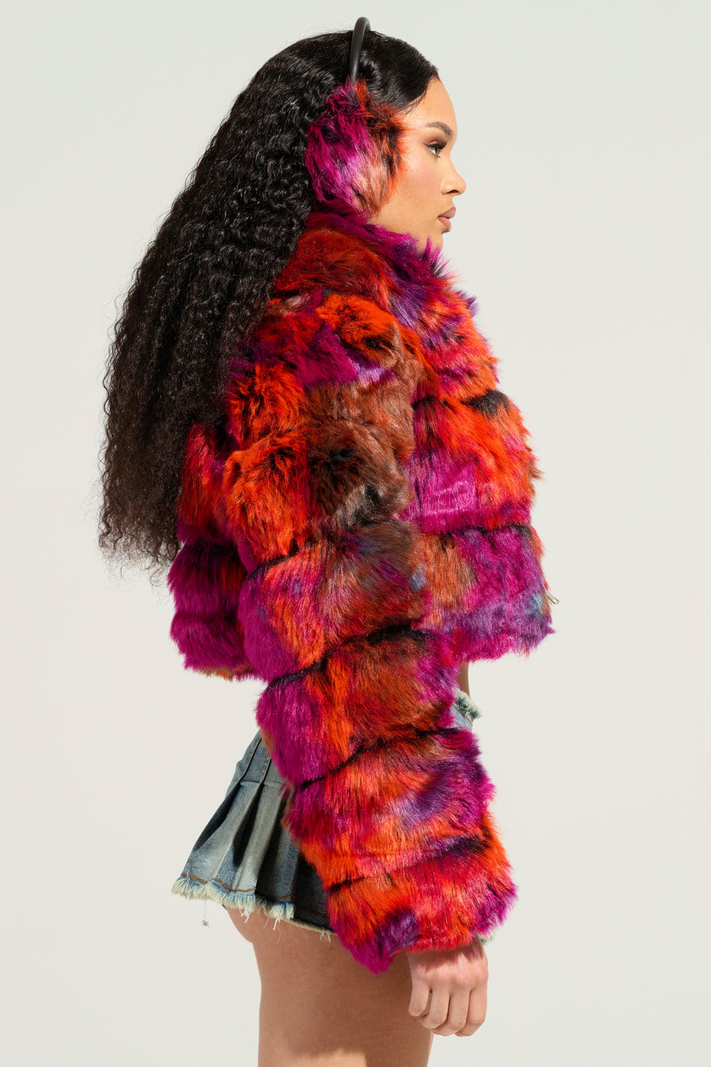 A person with long curly hair stands in profile, wearing the vibrant GRACELLE MULTI FAUX FUR CROPPED JACKET that showcases shades of red, orange, and purple. They pair it with black earmuffs and a short denim skirt against a plain background. 5