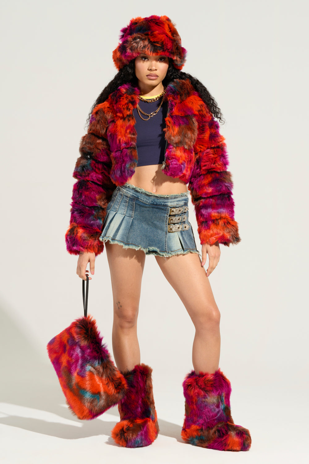 A person poses confidently wearing the GRACELLE MULTI FAUX FUR CROPPED JACKET, along with a matching hat and boots. They style it with a denim mini skirt, a navy cropped jacket, and hold a coordinating faux fur handbag against a plain, pale background. 8