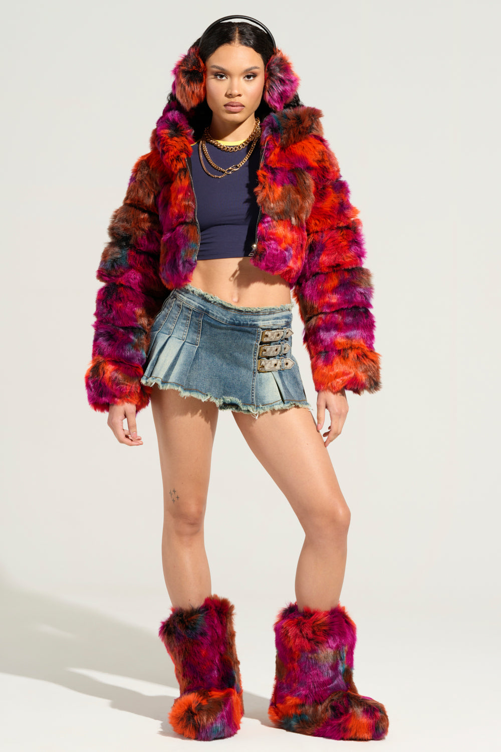 A person stands confidently in the GRACELLE MULTI FAUX FUR CROPPED JACKET, complemented by matching earmuffs and boots. They pair it with a navy crop top and a denim mini skirt featuring buckle details, set against a simple, light-colored studio backdrop. 9