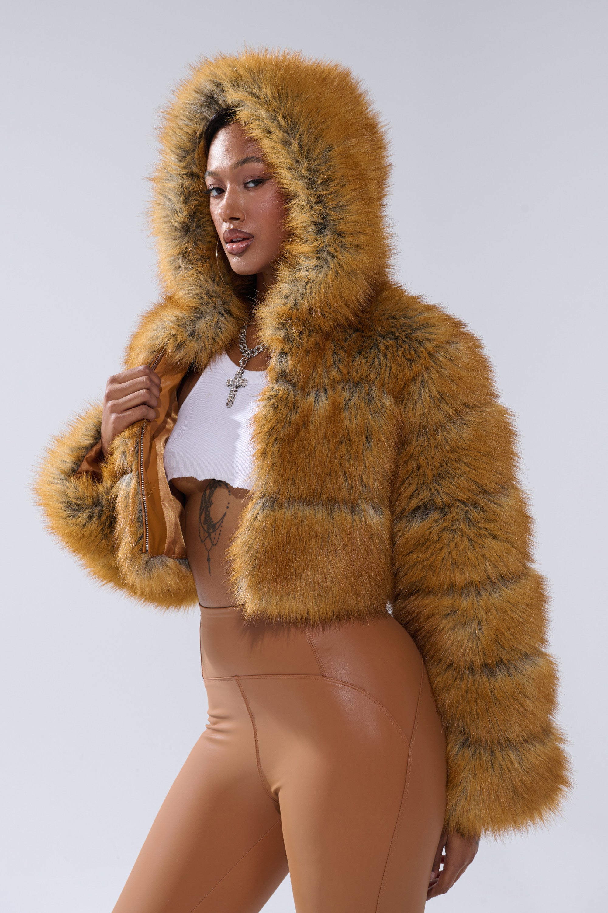 A woman confidently poses in the BREAK THE BANK HOODED FAUX FUR jacket, paired with tan high-waisted pants, a white crop top, and a cross necklace against a light gray background, holding the jacket stylishly in one hand.