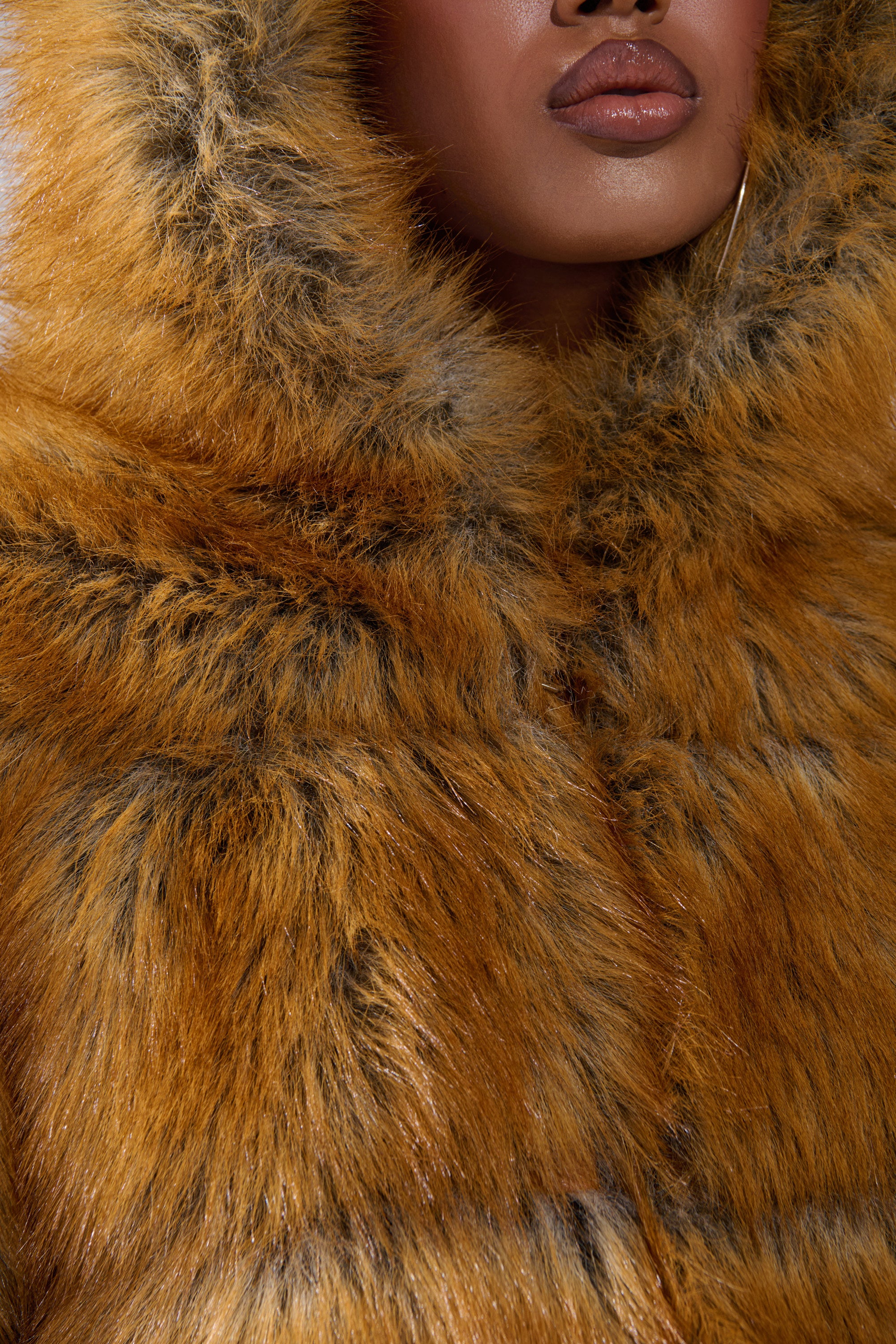A close-up shows a person in the BREAK THE BANK HOODED FAUX FUR coat in golden-brown, with only their glossy lips and brown skin visible beneath the plush hood.
