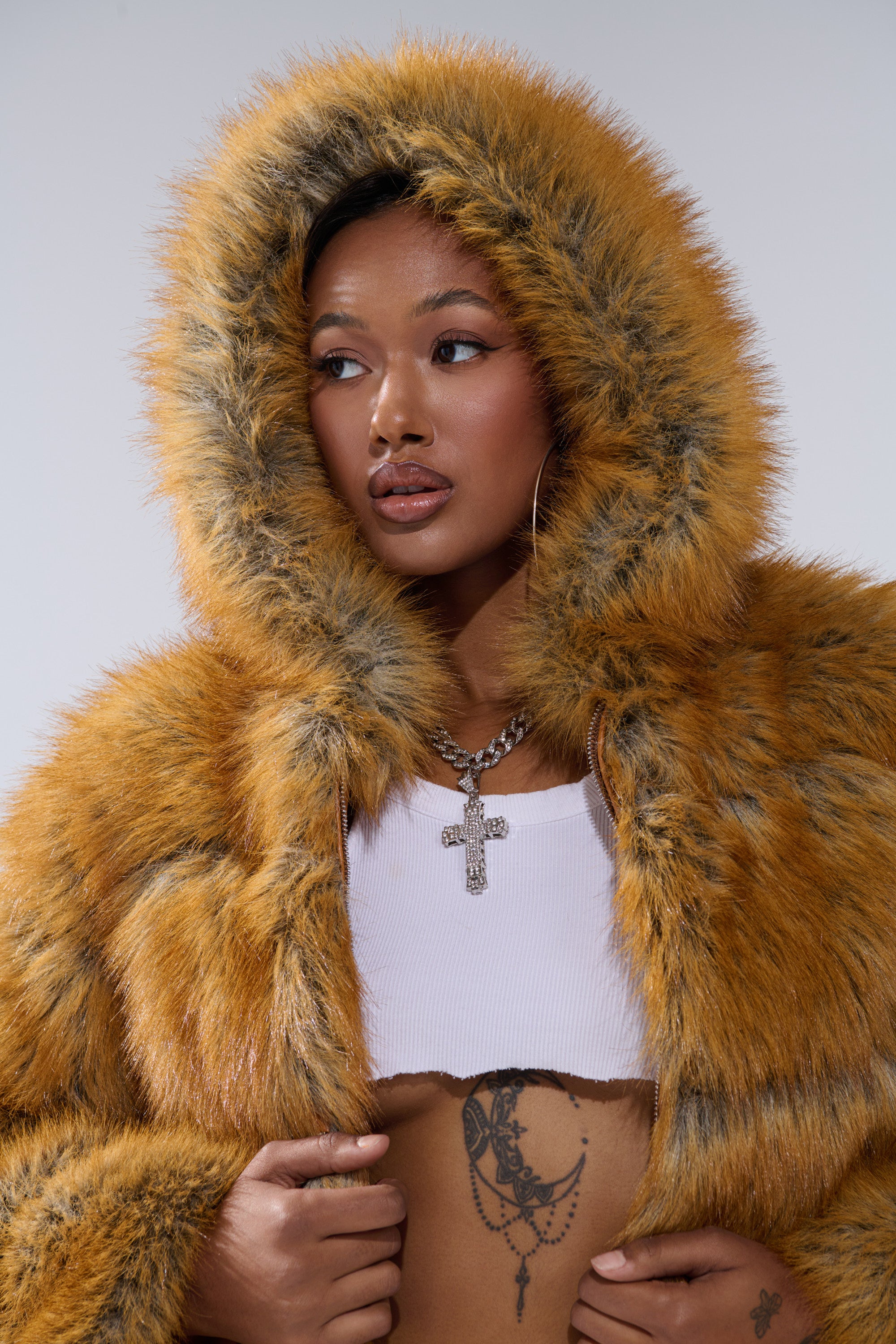 A woman in the BREAK THE BANK HOODED FAUX FUR jacket in bright orange with a white cropped top, cross necklace, stomach tattoo, and hoop earrings looks to the side against a neutral background.
