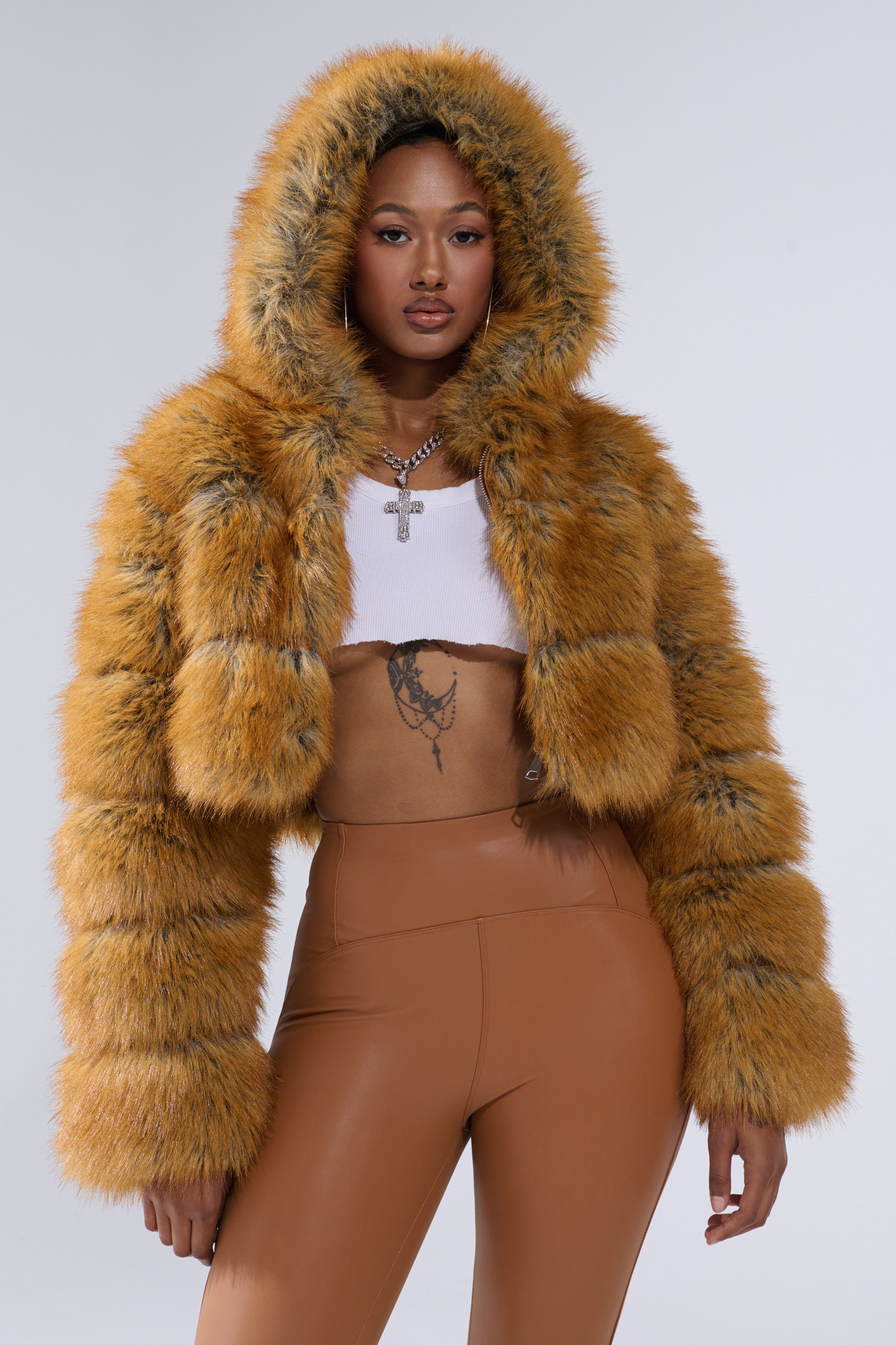 A woman exudes confidence in the BREAK THE BANK HOODED FAUX FUR jacket in vibrant orange, styled with brown high-waisted pants, a white crop top revealing her stomach tattoo, and a cross pendant chain necklace.