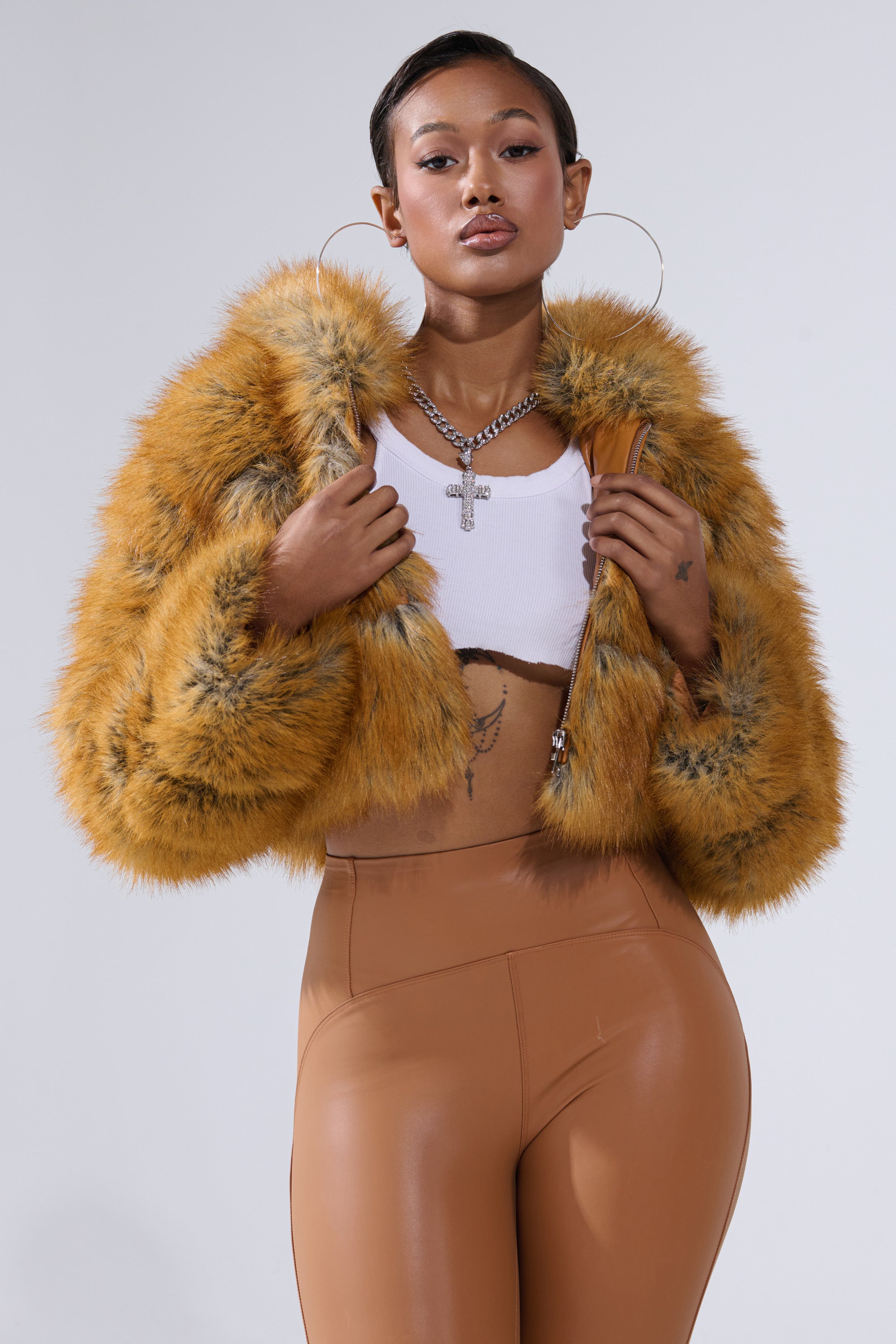 A woman poses confidently in the BREAK THE BANK HOODED FAUX FUR jacket, paired with a white cropped tank top and high-waisted caramel pants. She accessorizes with hoop earrings, a chain necklace, and displays visible tattoos.