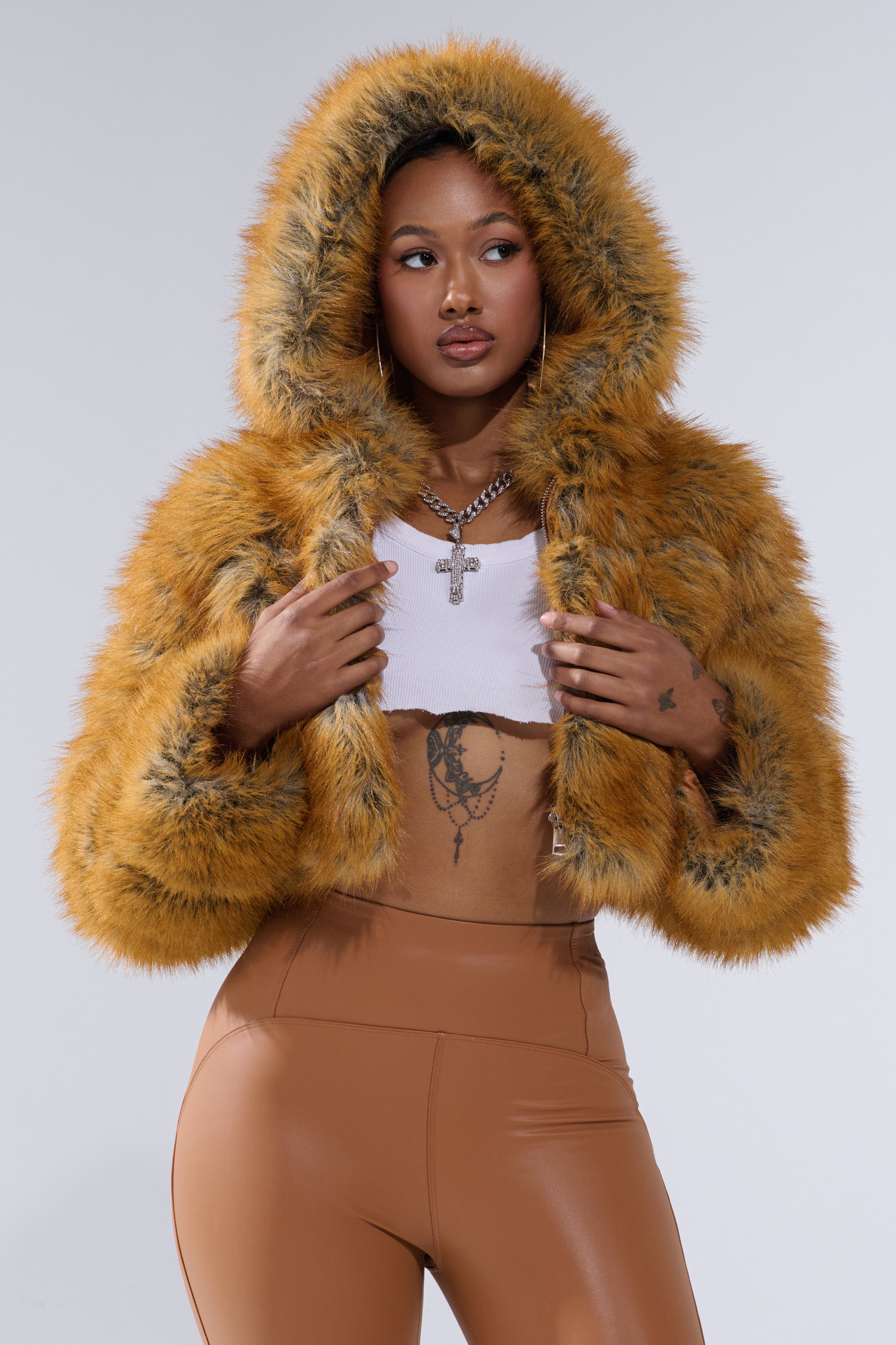 A woman confidently poses in the BREAK THE BANK HOODED FAUX FUR jacket in mustard yellow, styled with a white crop top, tan high-waisted pants, hoop earrings, layered necklaces, and visible tattoos on her chest and abdomen.