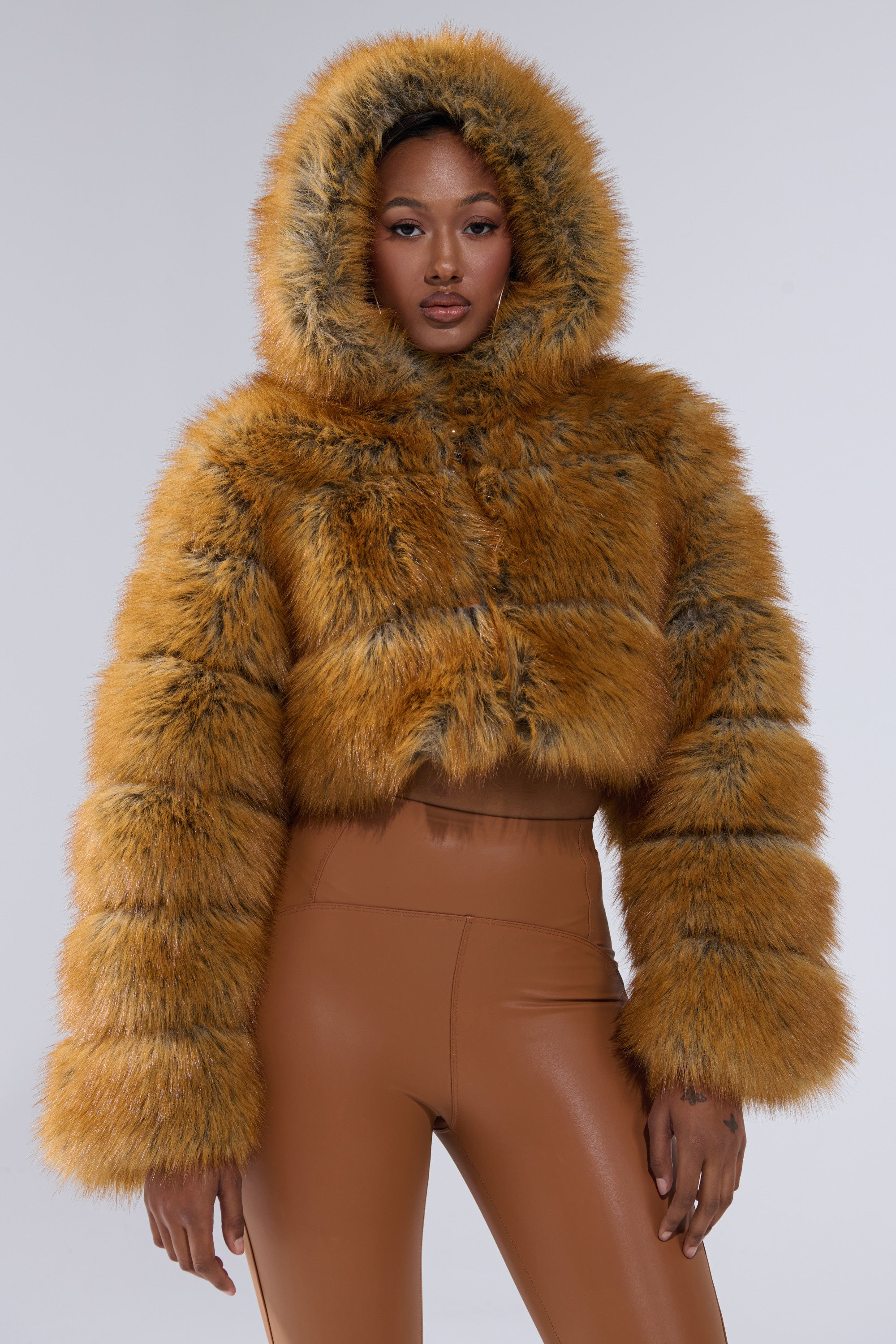 A woman confidently poses against a neutral background in the BREAK THE BANK HOODED FAUX FUR jacket, featuring a cropped fit in rich golden-brown, paired with high-waisted tan leather pants.