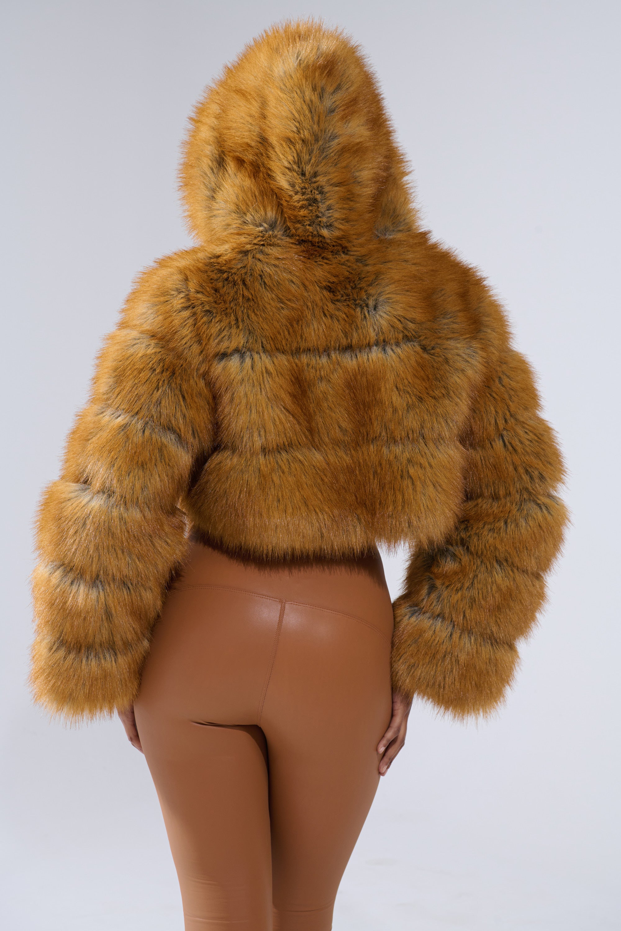 A person is seen from the back wearing the BREAK THE BANK HOODED FAUX FUR jacket in golden brown, cropped fit, with high-waisted shiny tan pants against a plain light background.