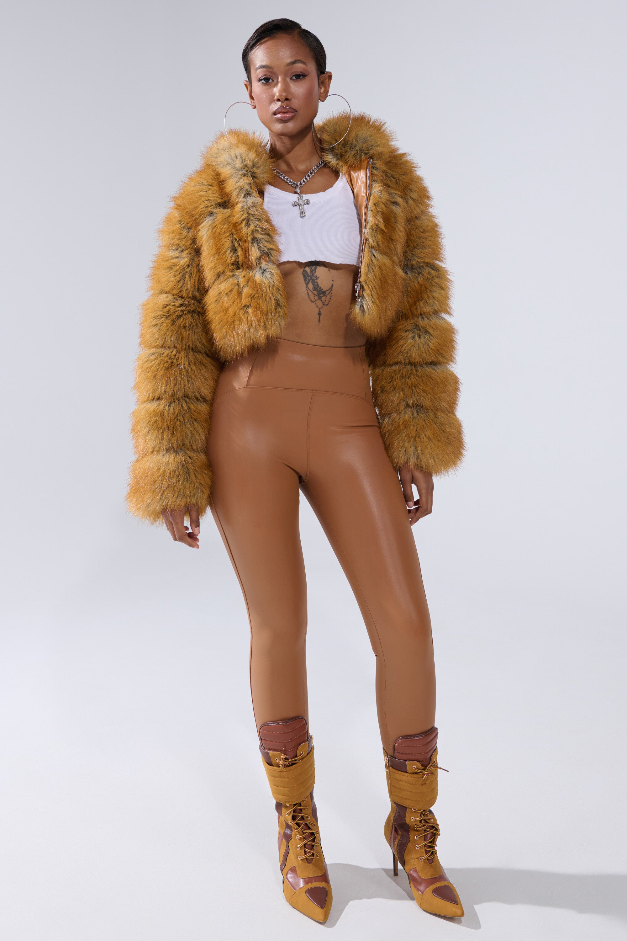 A woman stands confidently in the BREAK THE BANK HOODED FAUX FUR jacket with a cropped fit, paired with a white crop top, high-waisted tan leggings, tan lace-up heeled boots, large hoop earrings, and layered necklaces.