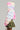 Dressed in the COTTON CANDY SKIES MULTI FAUX FUR BOMBER, a person radiates vibrant colors reminiscent of dreamy skies with its swirling pink, blue, and yellow hues. The bomber features a large hood and is paired with a green camouflage-patterned skirt adorned with playful ruffles. The simple gray background perfectly highlights their striking outfit.