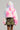 Wearing the COTTON CANDY SKIES MULTI FAUX FUR BOMBER, a person stands facing away. They're also in a camouflage mini skirt and holding a white bag with black dots. The plain gray background enhances the striking contrast of their vibrant attire.