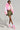 Dressed in the COTTON CANDY SKIES MULTI FAUX FUR BOMBER, a white crop top, camouflage mini skirt, and fuzzy pink and white boots, a person poses effortlessly with a studded handbag against a gray background reminiscent of cotton candy skies.
