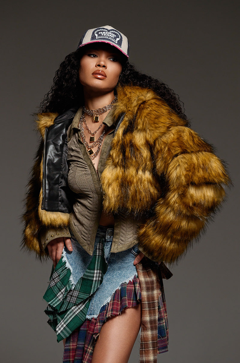 A person with curly hair confidently wears the GRACELLE Faux Fur Cropped Jacket, paired with a patchwork skirt and a green shirt. Layered necklaces and a trucker hat complete the look as they pose boldly against the plain background, blending style effortlessly with boldness.