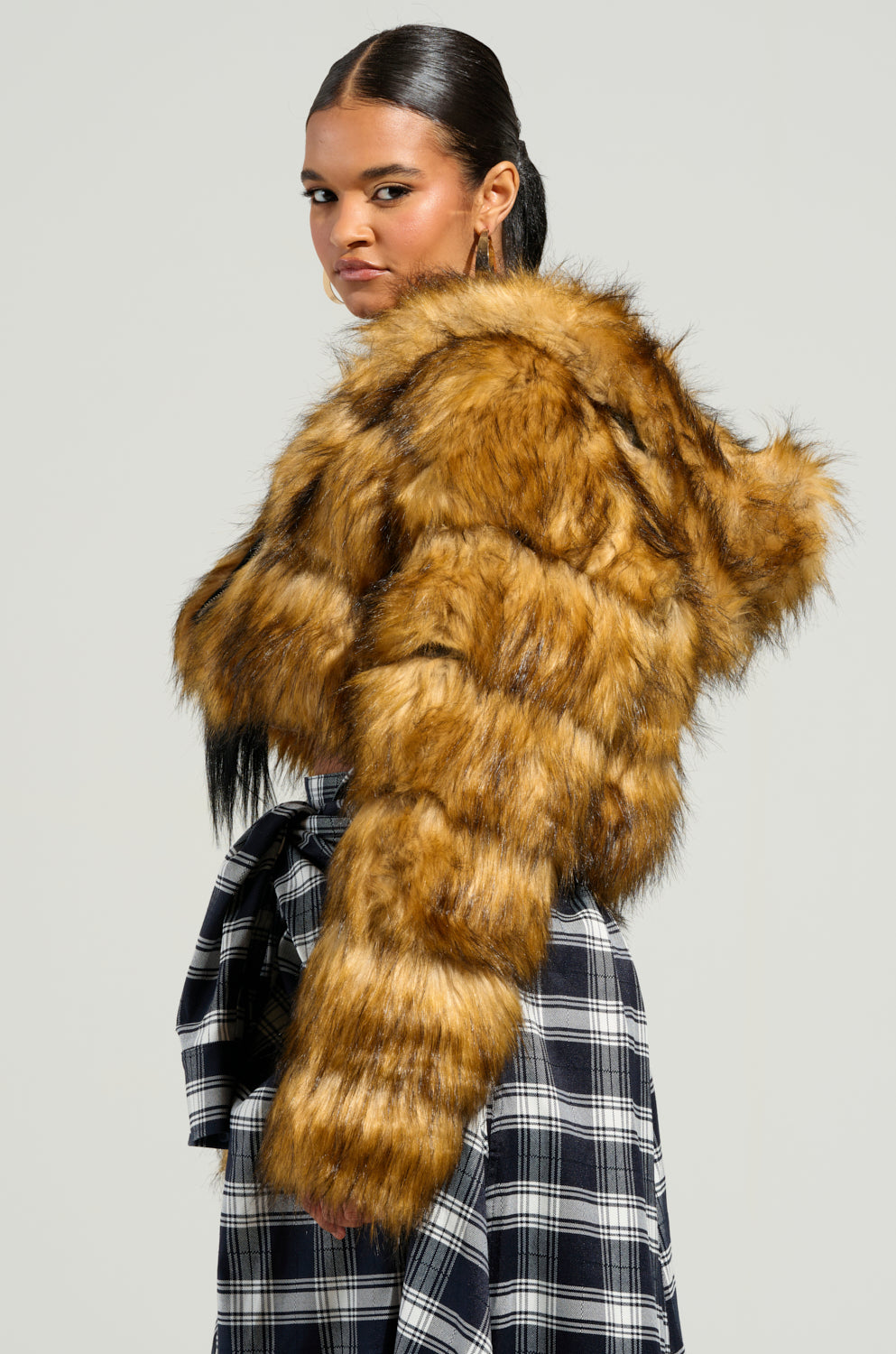 A person shows off a GRACELLE FAUX FUR CROPPED JACKET, stylishly paired with a black and white plaid skirt, radiating confidence. The jacket comes with a hood, and their hair is slicked back. Against a plain background, this chic ensemble is perfectly accentuated.