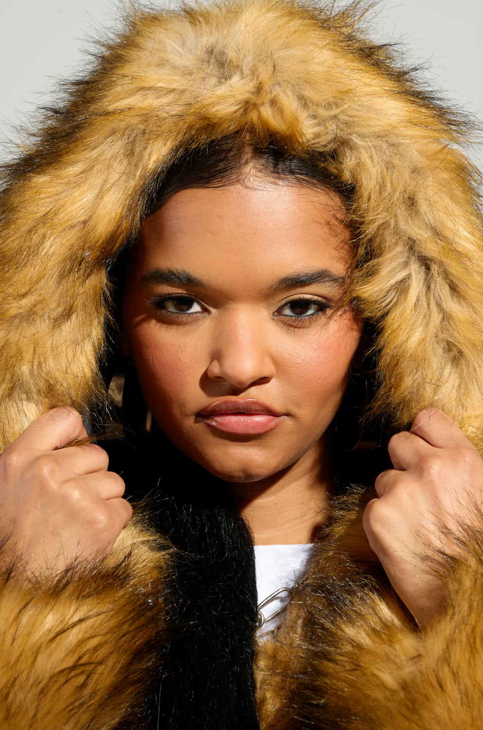 A person wearing a GRACELLE FAUX FUR CROPPED JACKET gazes directly at the camera. This jacket, which includes a fur-lined hood and showcases a mostly brown color with black accents, is being worn with the individual's hands raised, holding the edges of the hood against a plain, light-colored background.