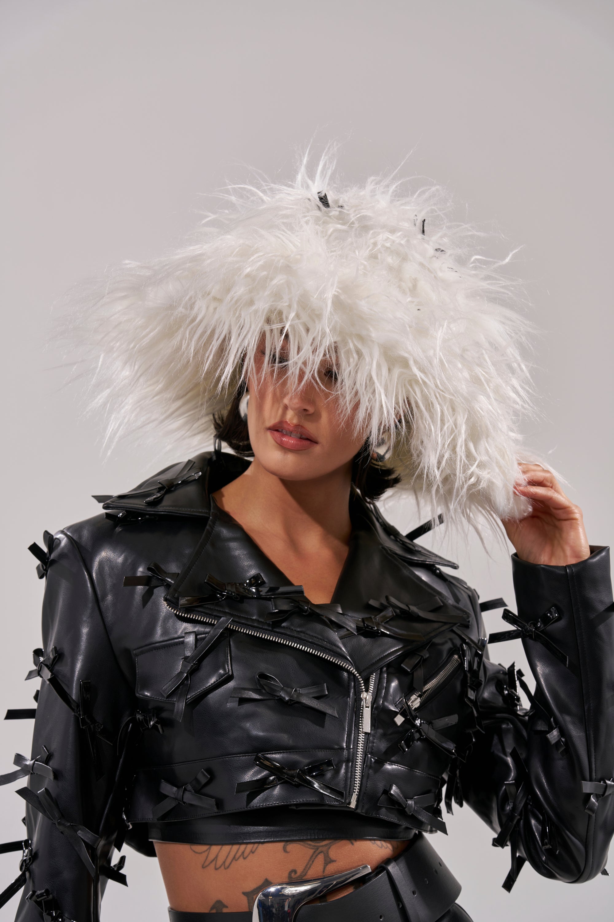 A person wearing the WITH A BOW FAUX FUR BUCKET HAT and a black cropped leather jacket adorned with satin bows stands against a plain, light background. 0