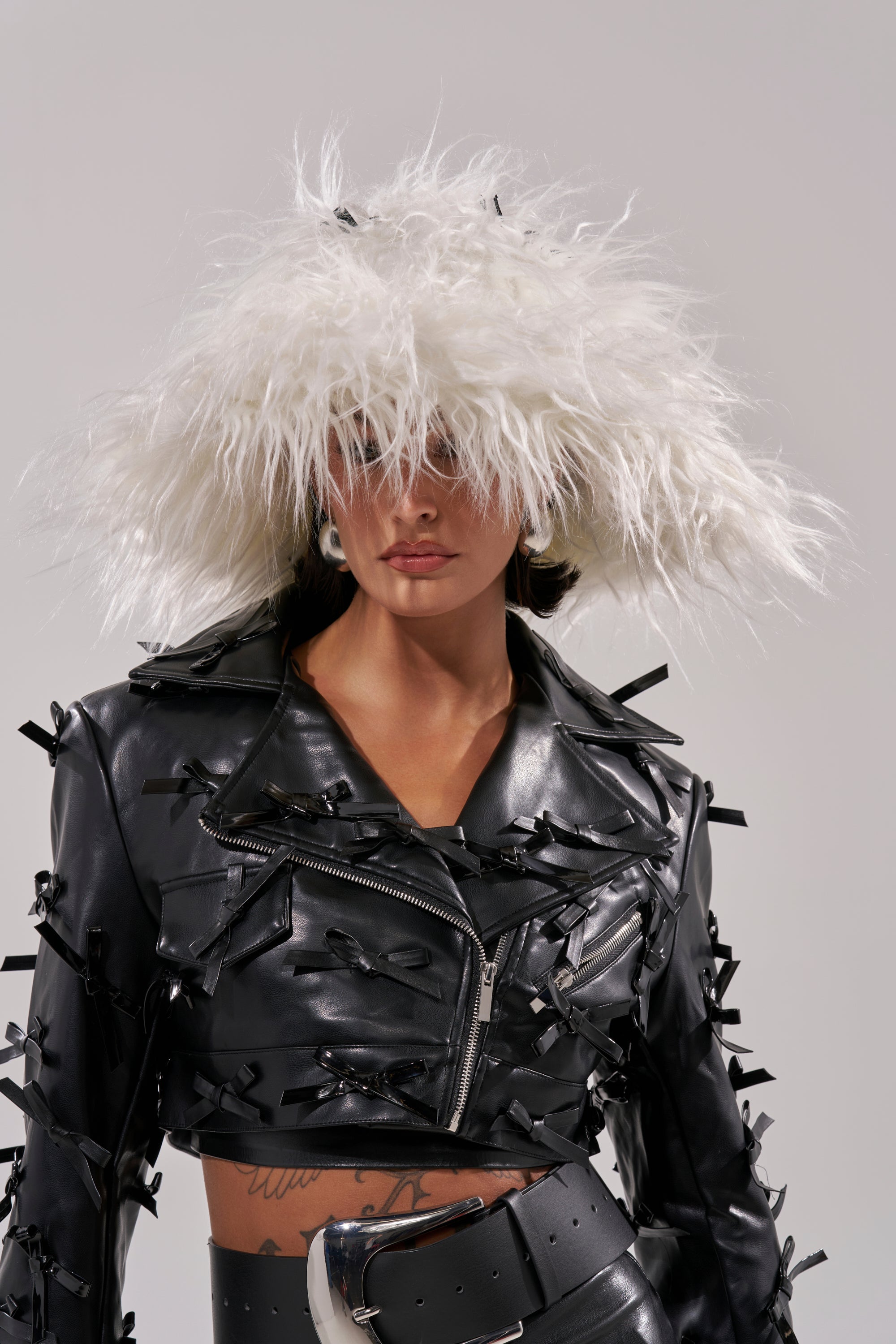 Wearing the WITH A BOW FAUX FUR BUCKET HAT and a black leather jacket with satin bows, this person stands against a plain background. The hat partially covers their eyes, adding dramatic, edgy flair to the coquette-inspired look. 1