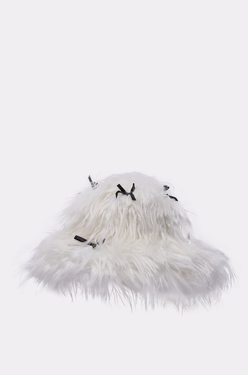 The WITH A BOW FAUX FUR BUCKET HAT is a white faux fur bucket hat with long, shaggy fibers and multiple small black satin bows, shown against a plain light background. 3