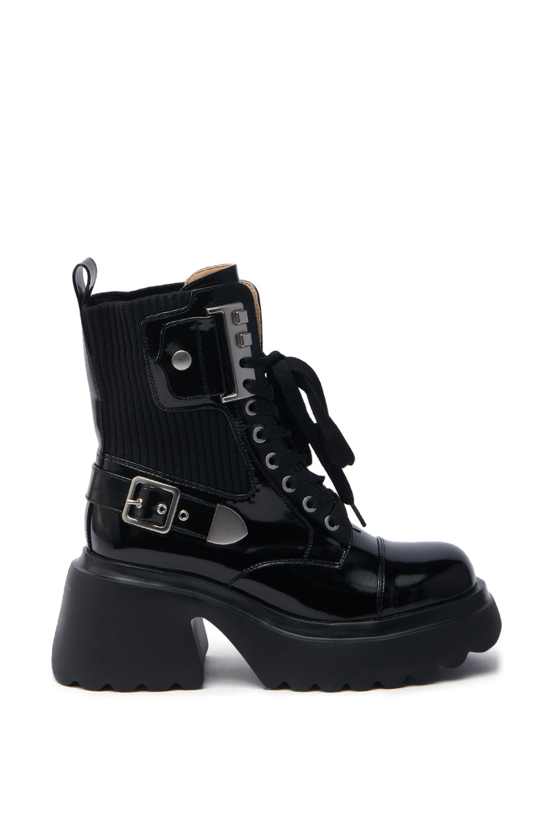 The AZALEA WANG GRAY BLACK WITH METAL PLATFORM BOOTIE features a shiny black faux leather design with laces, a side buckle, and ribbed fabric around the ankle. It has a thick platform heel with a chunky sole featuring a rugged pattern and silver hardware details.