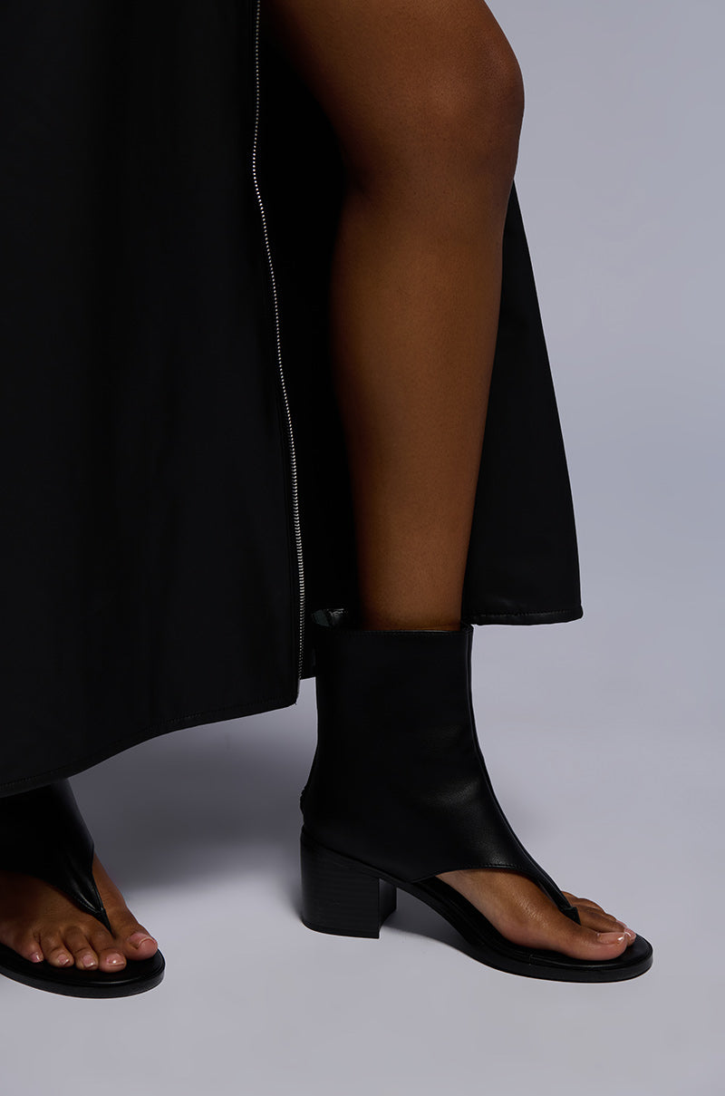 A person stands on a light gray background wearing the AZALEA WANG GRAYSON BLACK CHUNKY GLADIATOR SANDAL with an open design and chunky heel. Part of their black outfit, including a long skirt or dress, is visible.