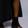 A person stands on a light gray background wearing the AZALEA WANG GRAYSON BLACK CHUNKY GLADIATOR SANDAL with an open design and chunky heel. Part of their black outfit, including a long skirt or dress, is visible.