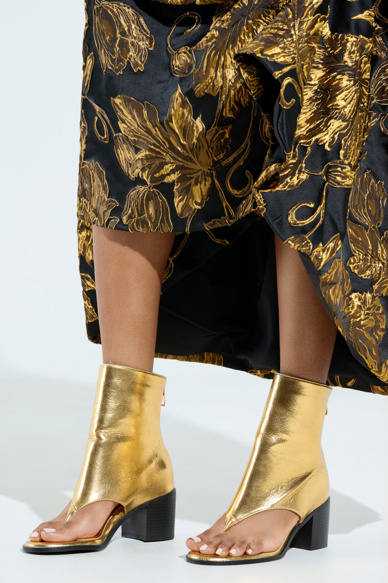 A model wears a black and gold floral dress paired with metallic AZALEA WANG GOLD CHUNKY SANDALS. The chunky heel complements the shiny texture, with the dress elegantly draping and partially covering these striking shoes.