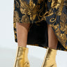 A model wears a black and gold floral dress paired with metallic AZALEA WANG GOLD CHUNKY SANDALS. The chunky heel complements the shiny texture, with the dress elegantly draping and partially covering these striking shoes.